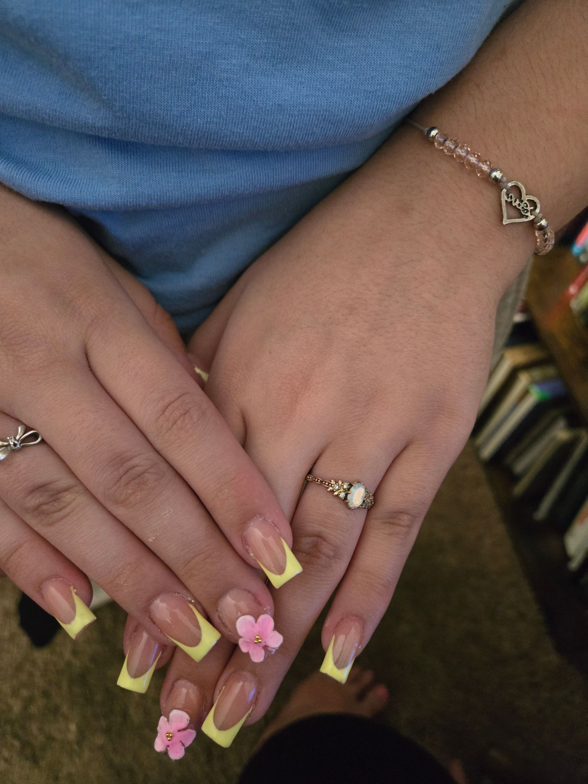 Hands with long pastel yellow and pink floral nails, wearing a blue sweater and silver jewelry.