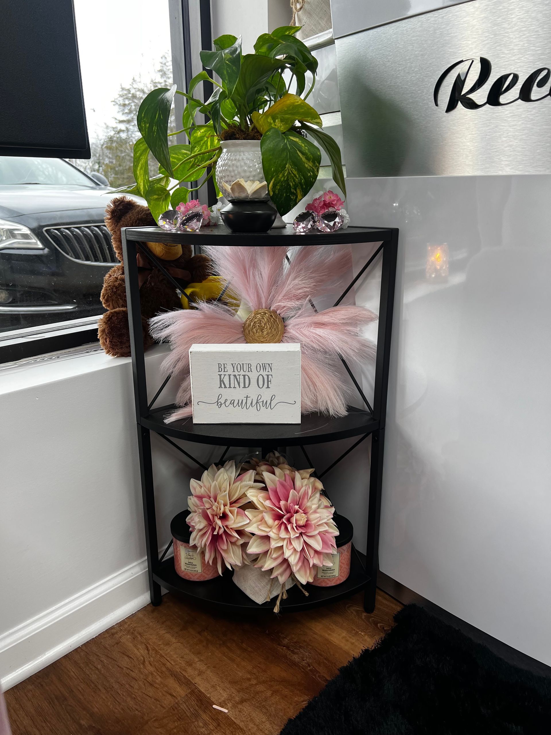 Floral display on a black shelf beside a window and white wall, with pink flowers and a small sign.
