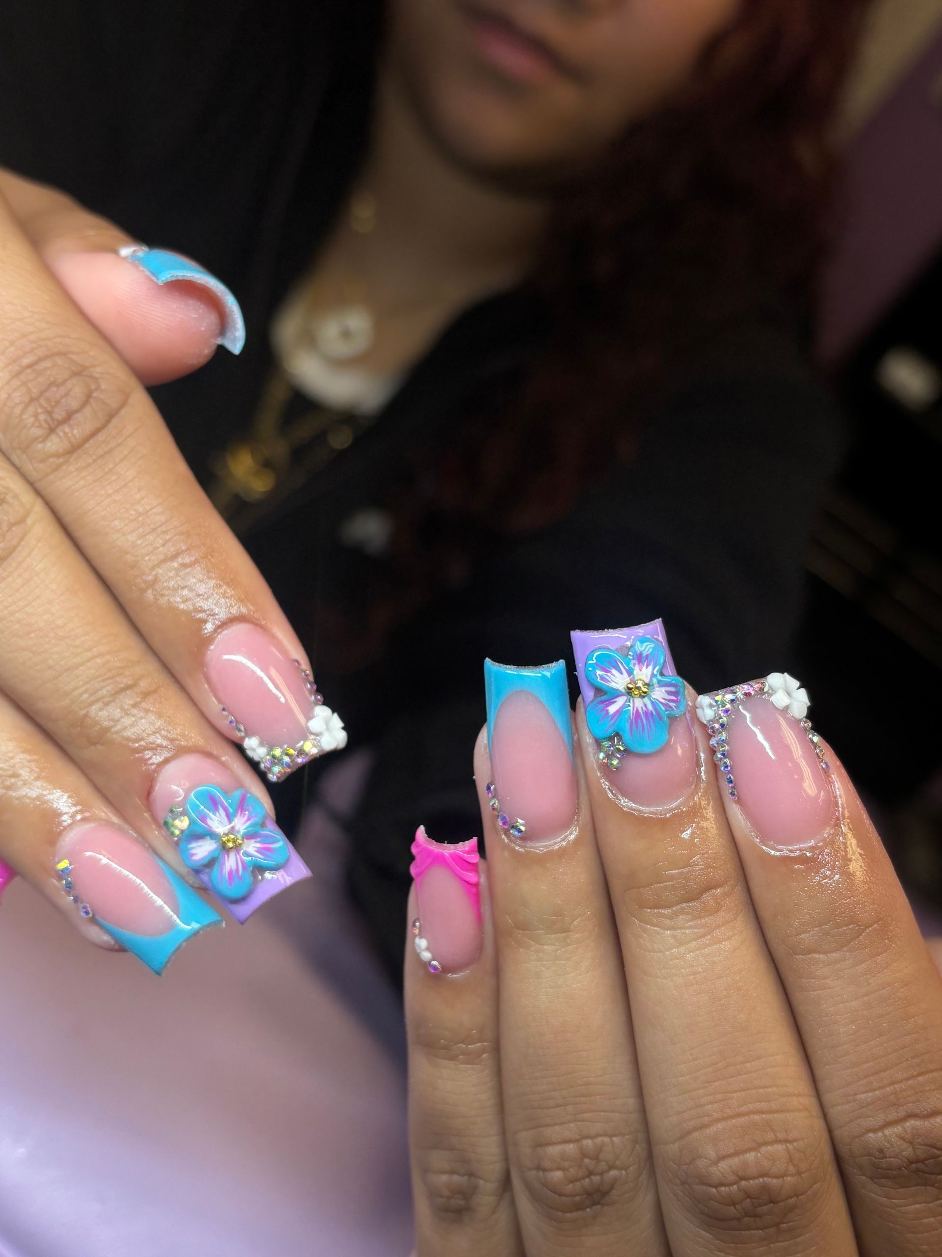 Close-up of hands showing long pink nails with blue tips, glitter, and floral rhinestone accents