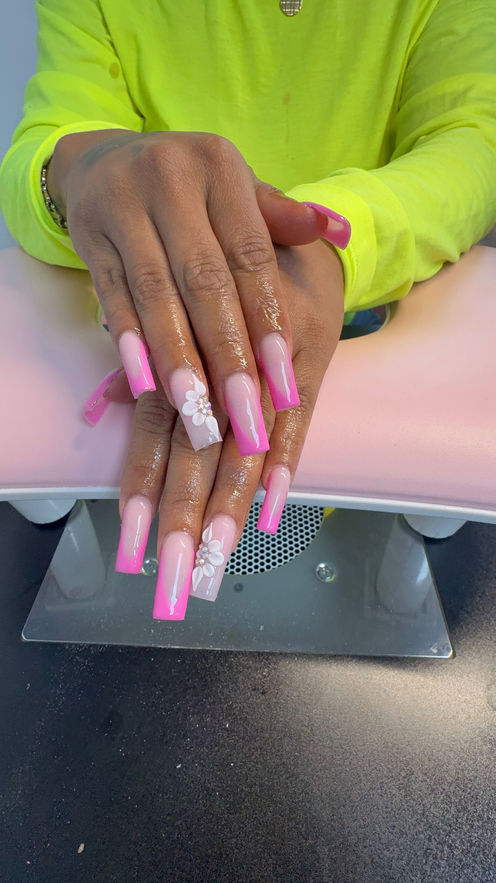 Manicured hands with long pink nails resting on a white armrest over a nail salon table