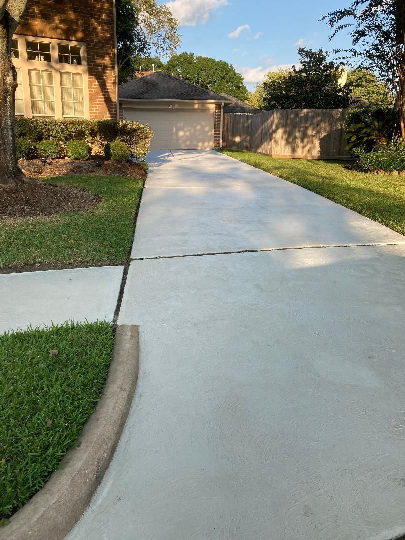 Residential driveway leading to a garage, bordered by grass, trees, and a brick house at sunset.