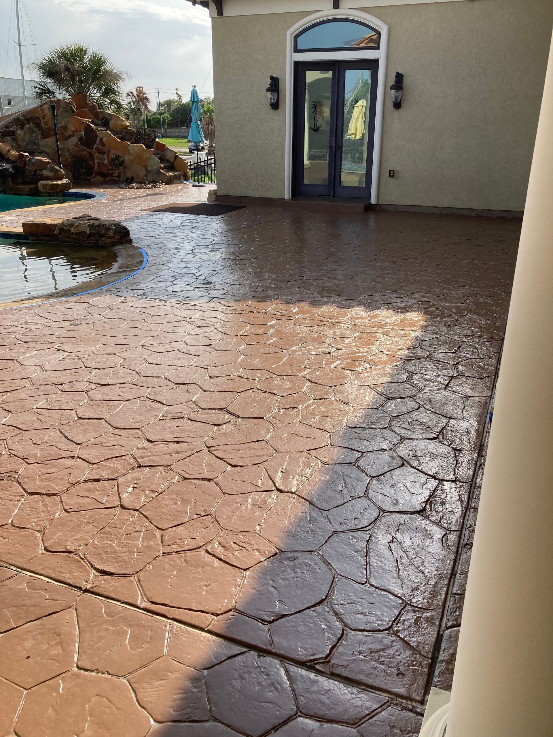 Wet stamped-concrete patio beside a house entrance, with puddles and bright sunlight.