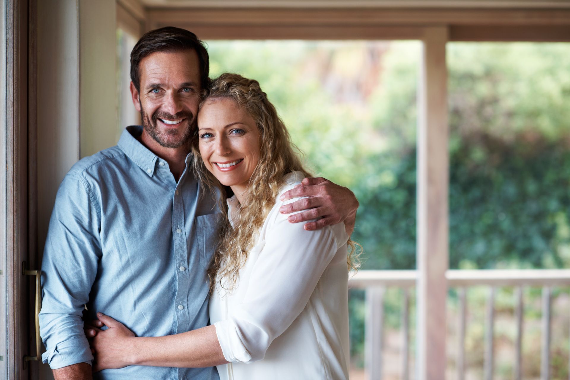 Smiling couple embracing in a bright doorway, with a porch and greenery in the background