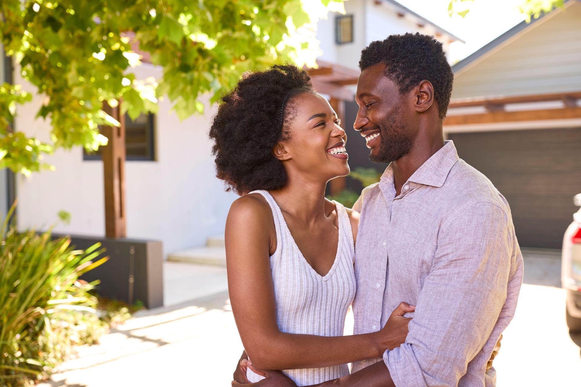 Two people smiling and embracing outdoors near a house, with sunlight and green leaves overhead