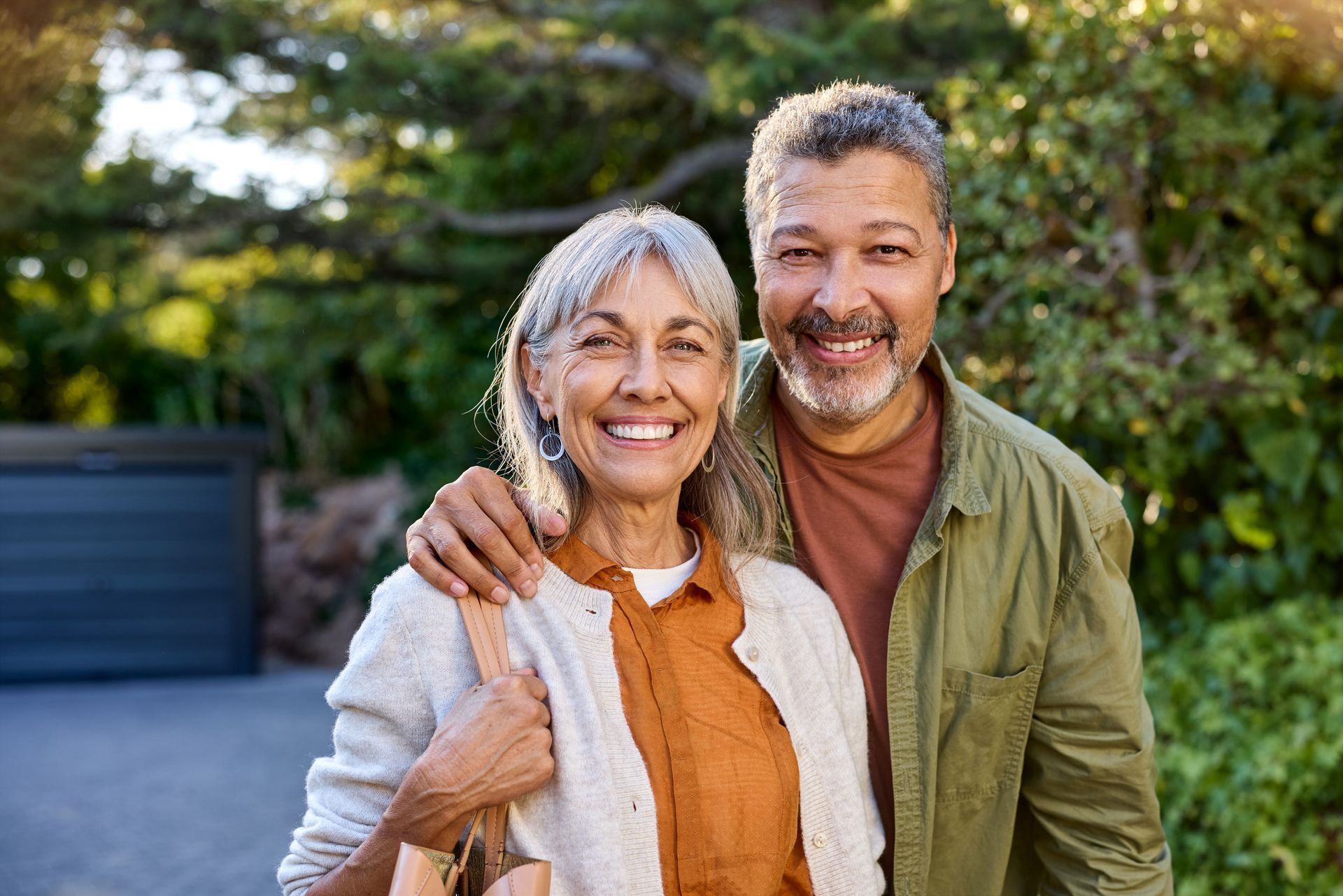 Two smiling people standing outdoors, one with an arm around the other in a garden.