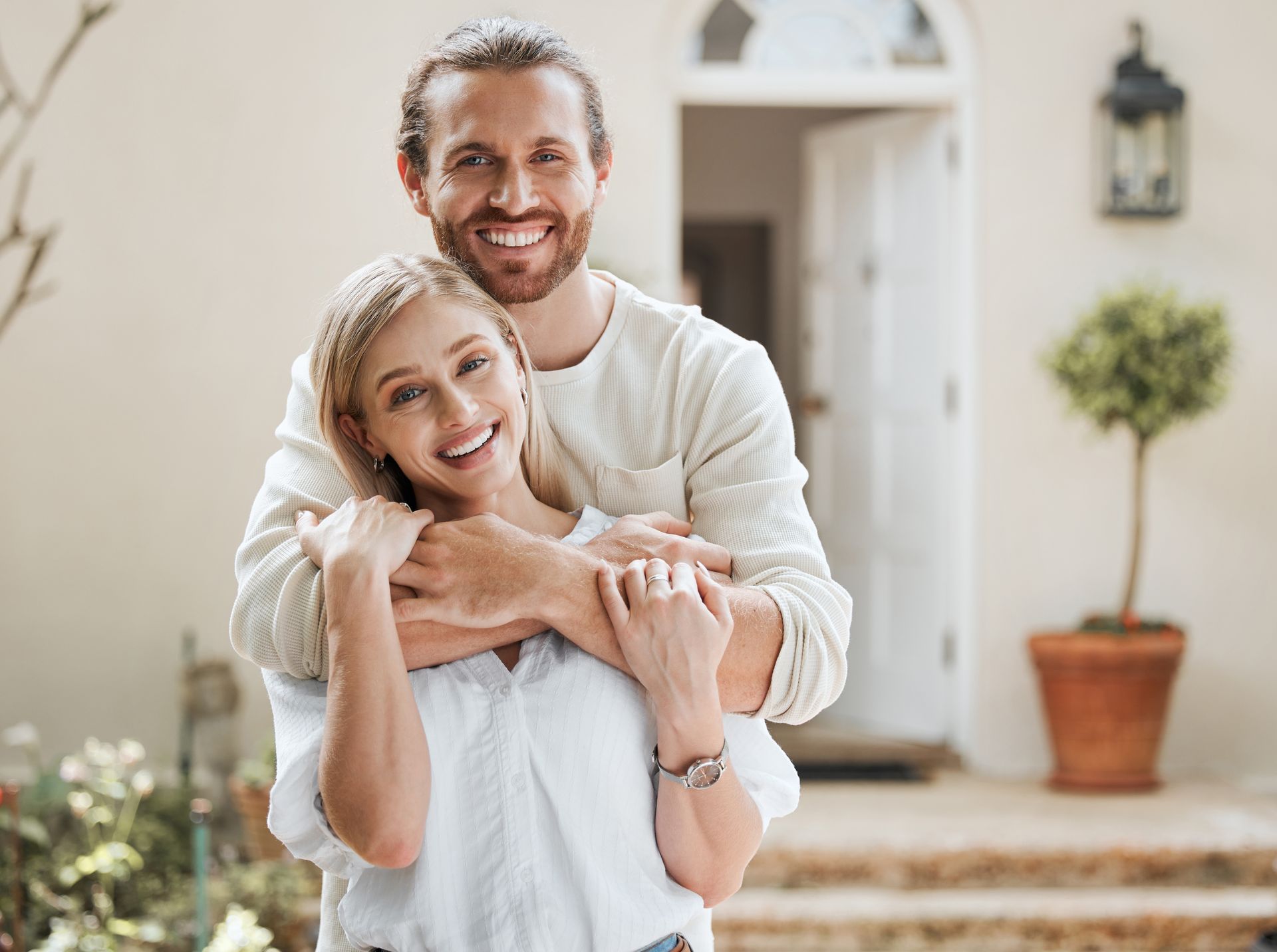 Smiling couple embracing on a sunlit patio by a white house and potted plants