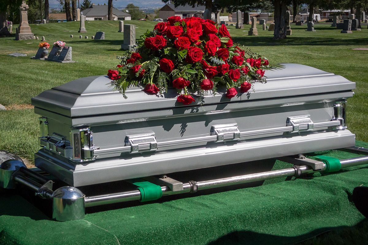 Silver casket with red rose arrangement, set on a green cloth in a cemetery.