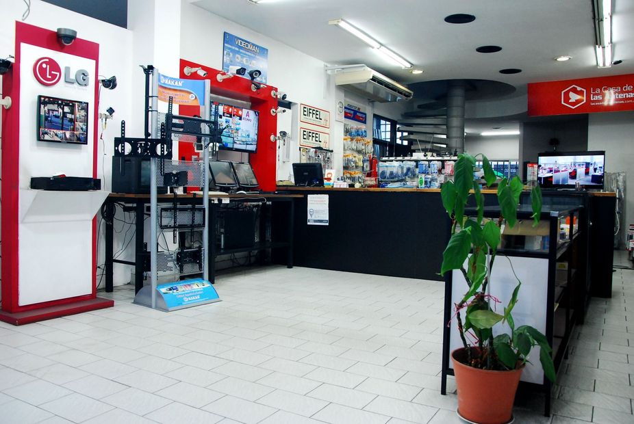 Interior luminoso de una tienda de electrónica con vitrinas rojas, televisores, mostradores y plantas en macetas.