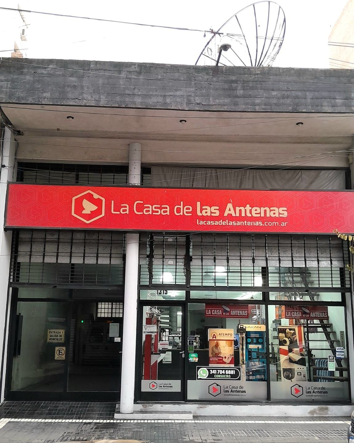 Fachada de “La Casa de las Antenas” con letrero rojo y entrada de cristal en una calle de la ciudad.
