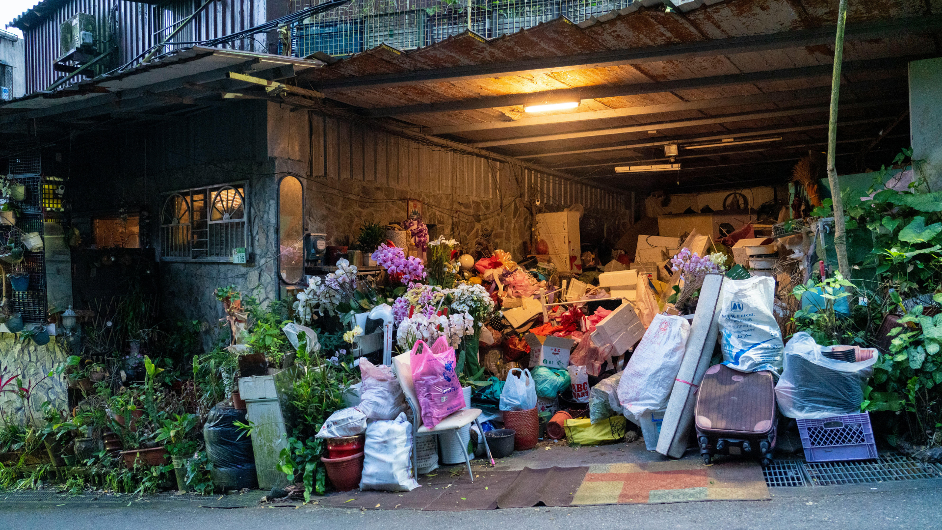 A cluttered outdoor storage area filled with flowers, bags, and miscellaneous items; dim lighting.