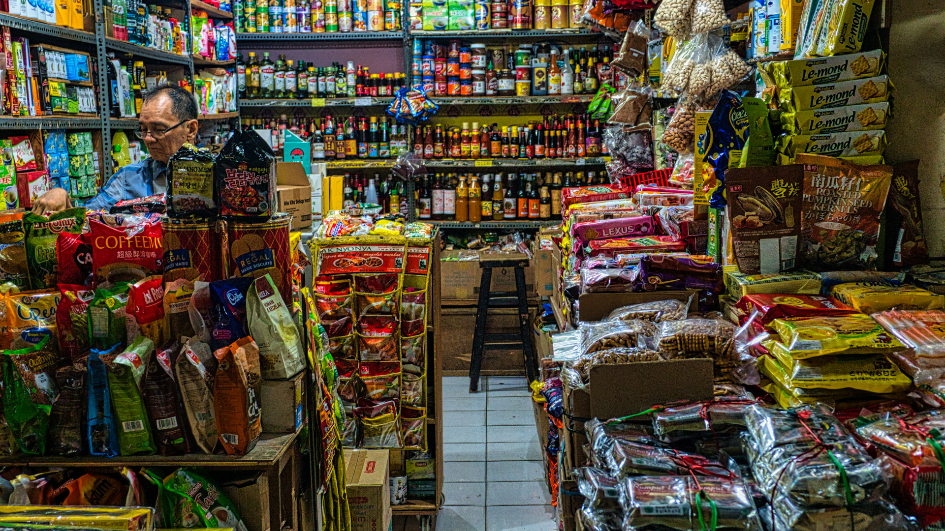 Small shop interior packed with various food items. A man stands behind a counter.