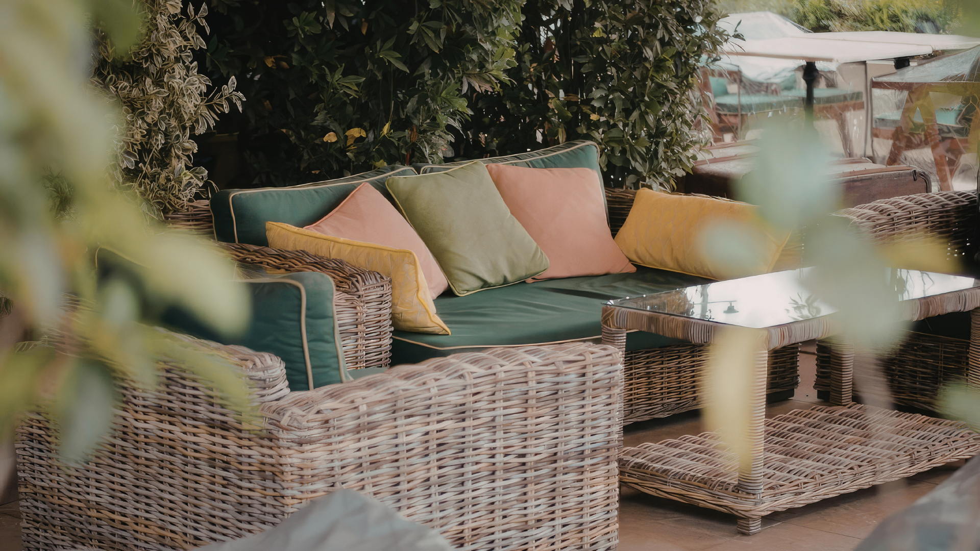 Outdoor wicker lounge set with green cushions and multicolored pillows on a patio, partially obscured by foliage.
