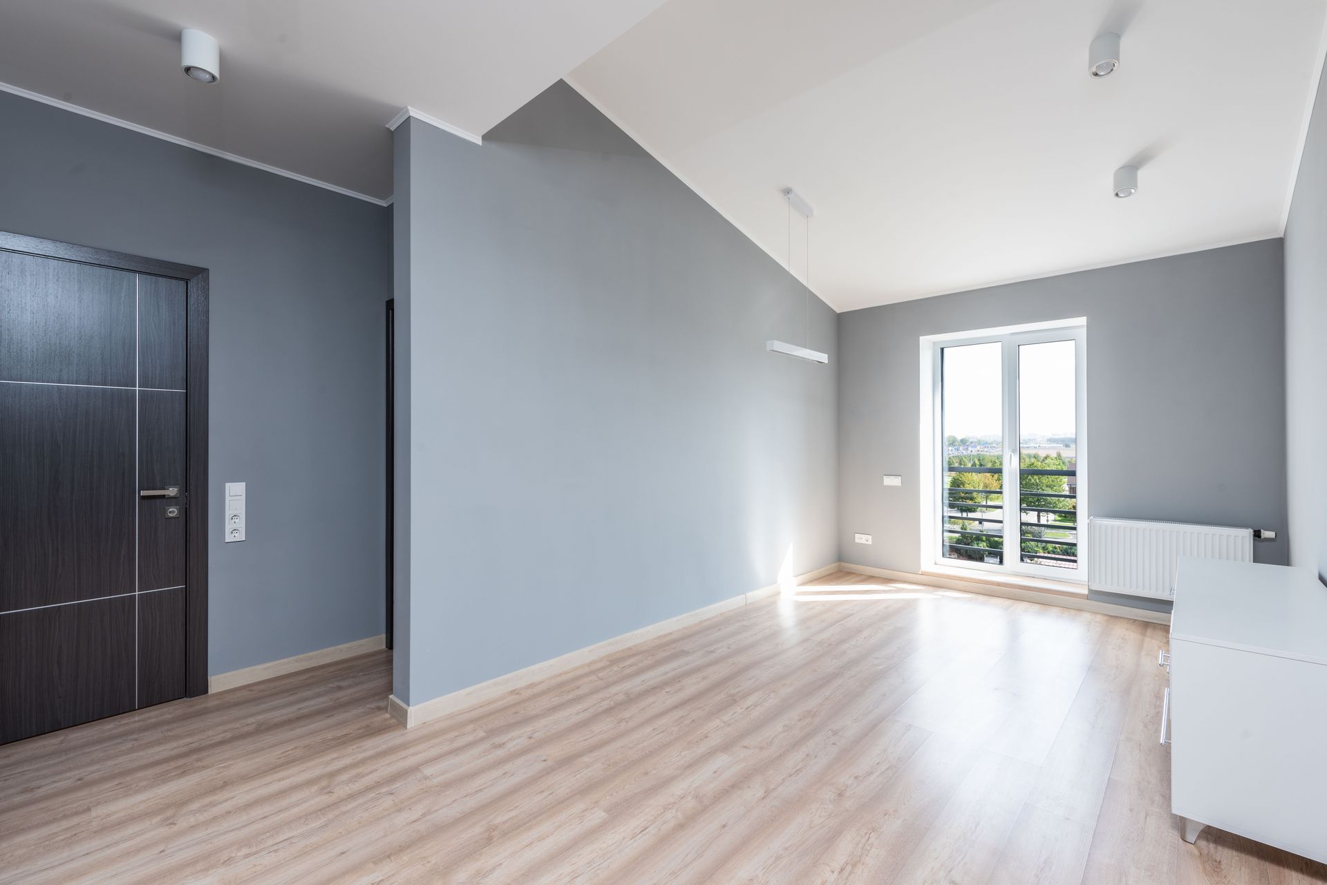 Empty modern apartment room with gray walls, light wood floors, and a bright windowed door to a balcony