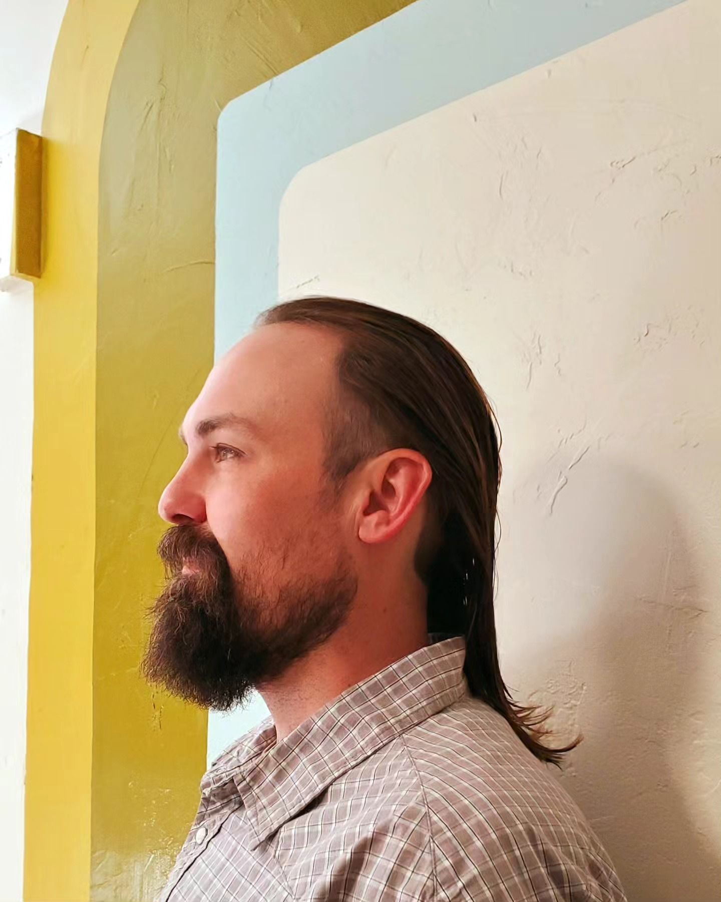 Side profile of a bearded man with tied-back hair standing against a pastel yellow and blue wall.