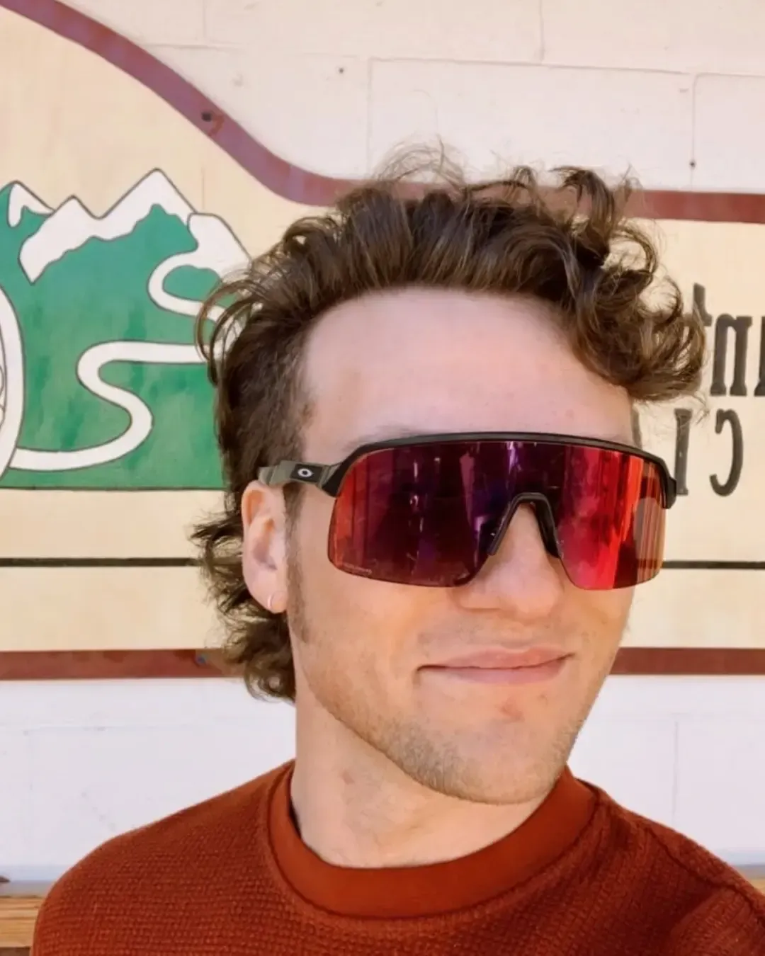 Person wearing red mirrored sunglasses and an orange sweater, standing indoors against a painted wall.