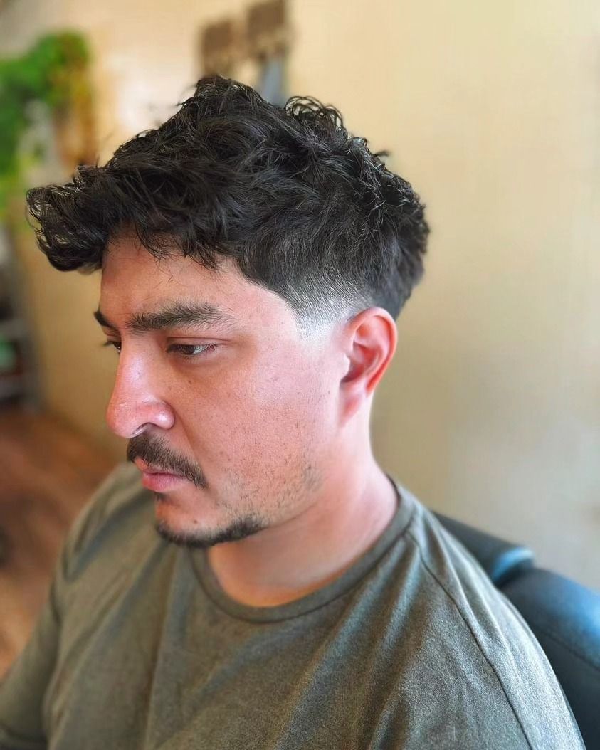 Side-profile portrait of a man with curly dark hair and a mustache, seated indoors against a blurred background