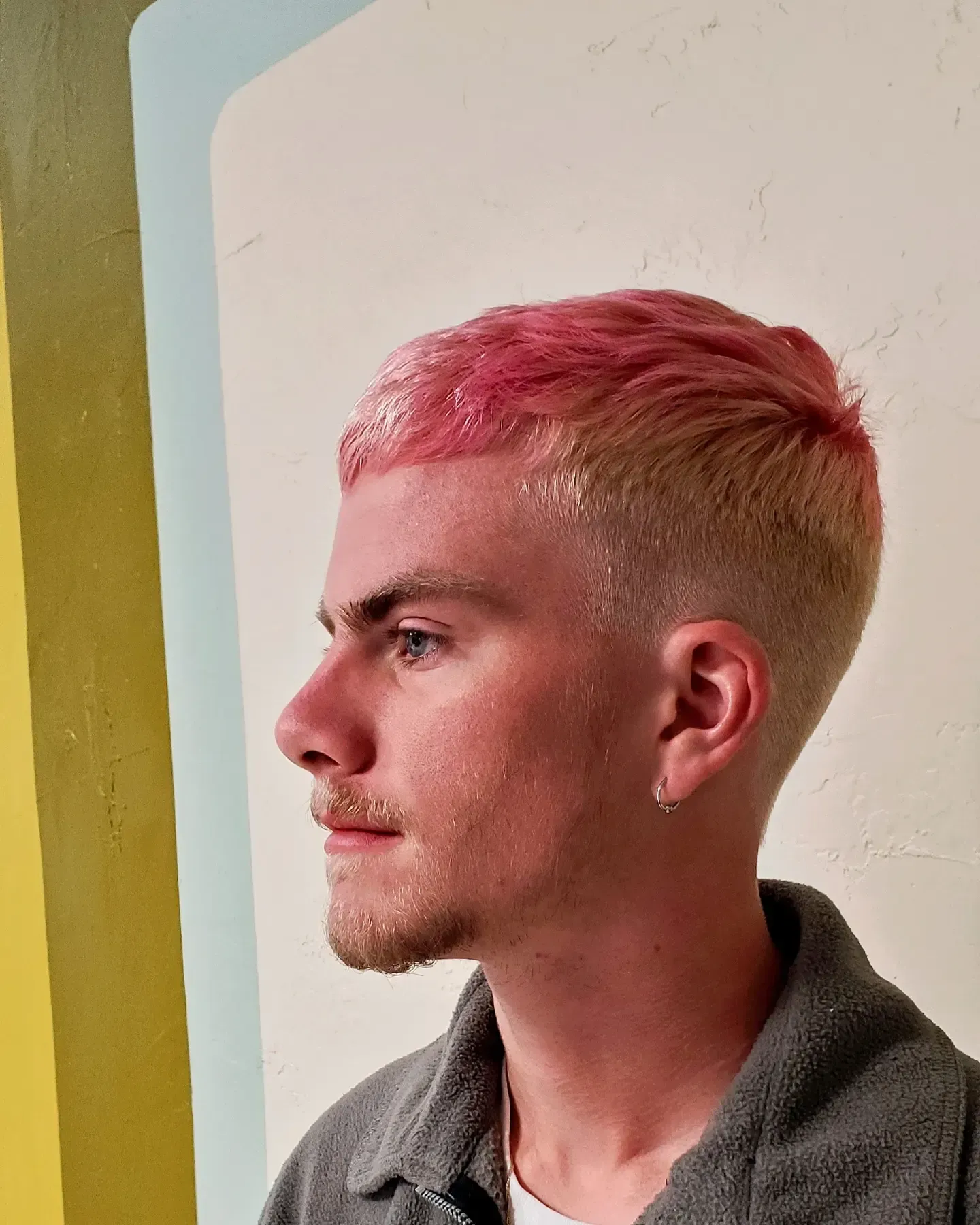Side profile of a person with short pink hair wearing a gray top against a light wall.