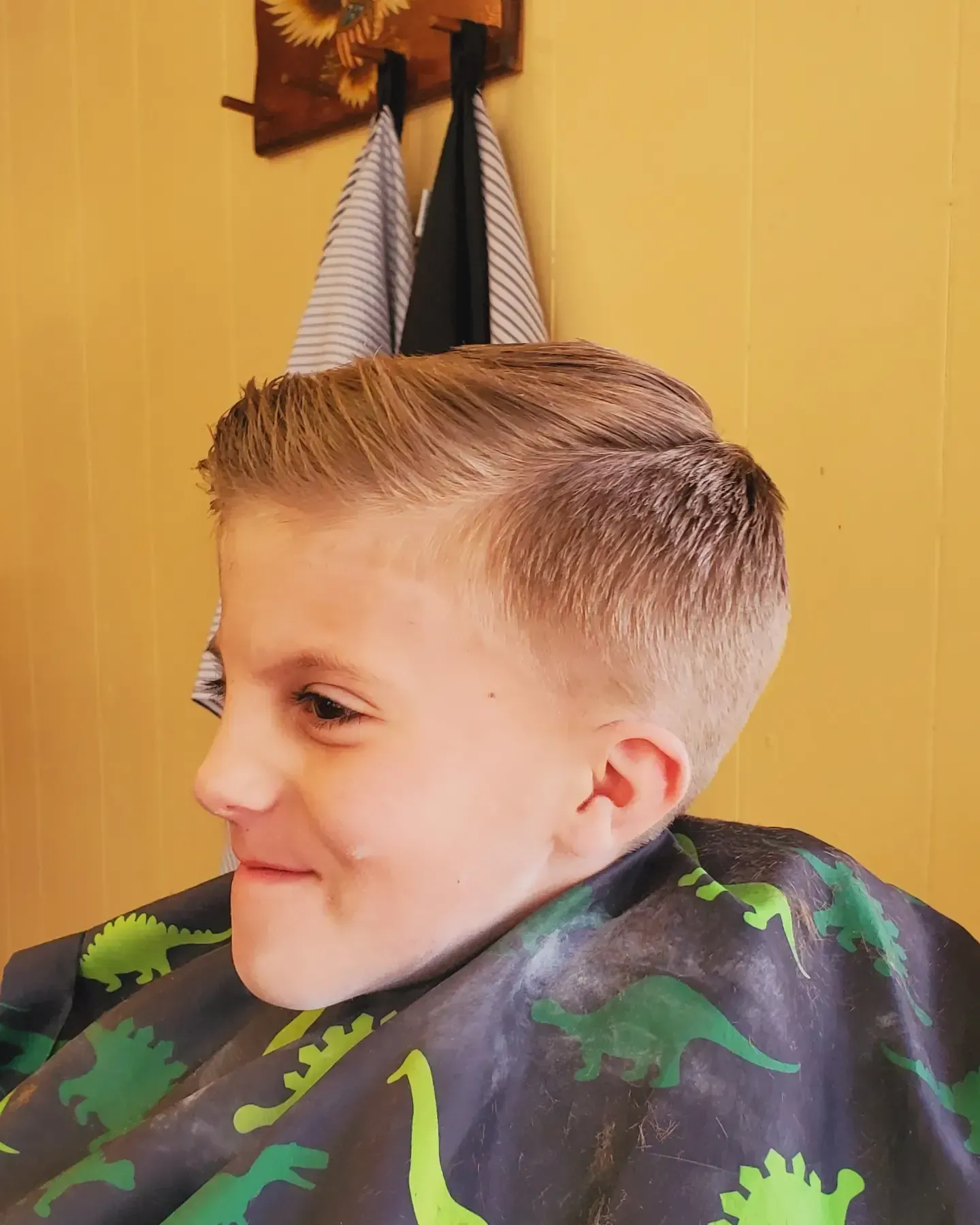 Profile view of a smiling child with a fresh haircut in a barber cape, indoors against a yellow wall
