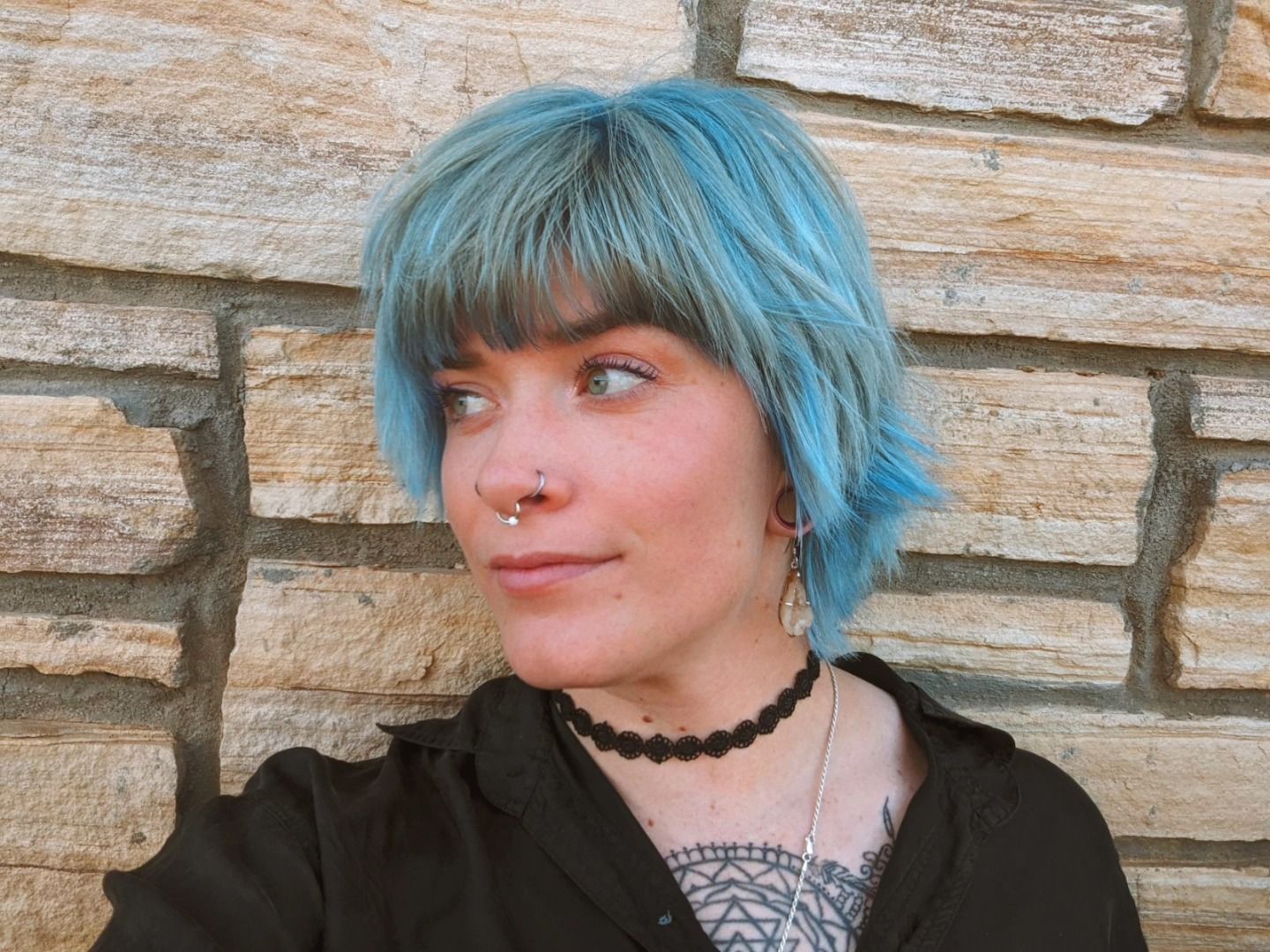 Portrait of a person with teal hair and a black choker, standing before a stone wall, looking left