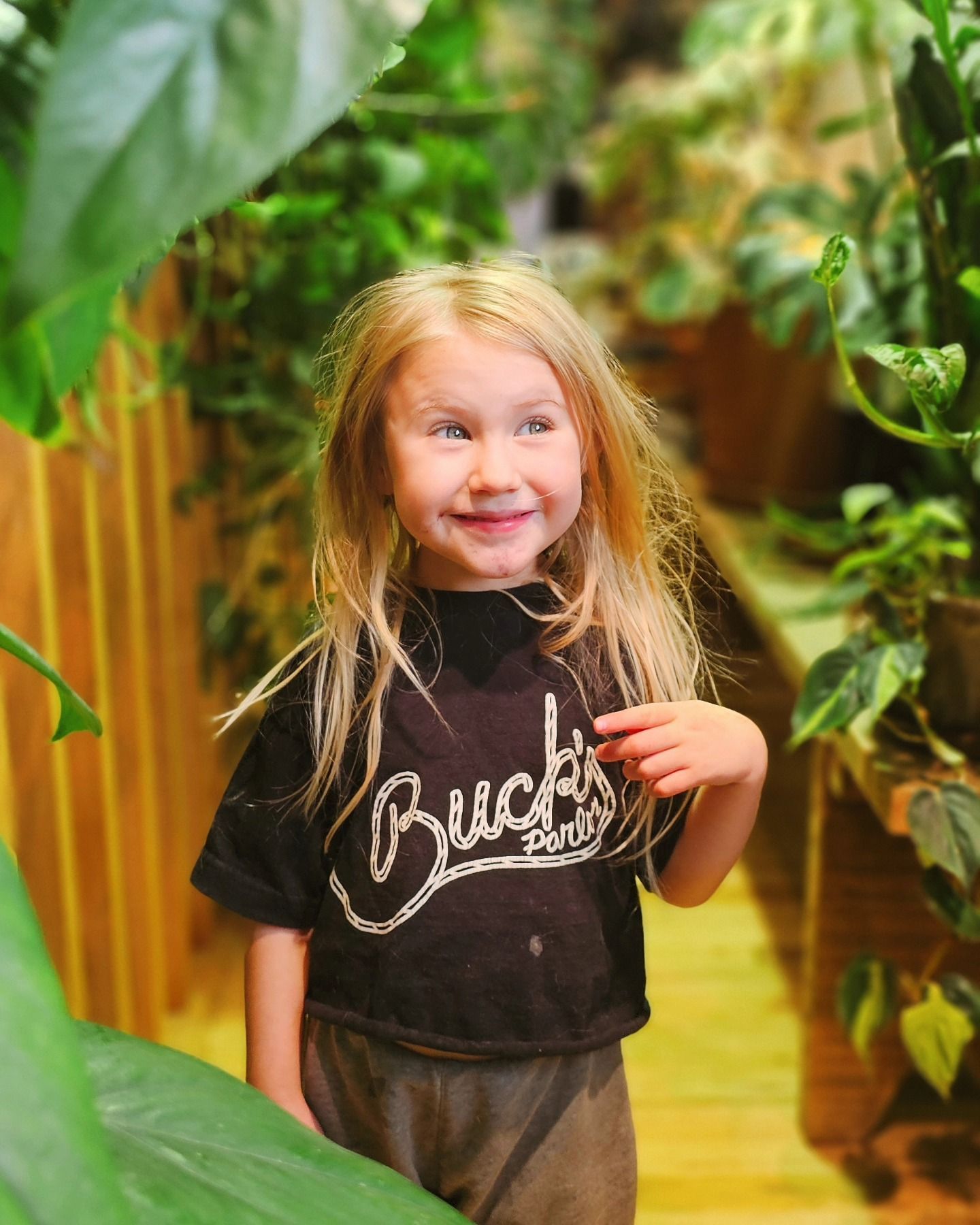 Smiling child in a black shirt standing among lush green plants in a bright indoor garden path
