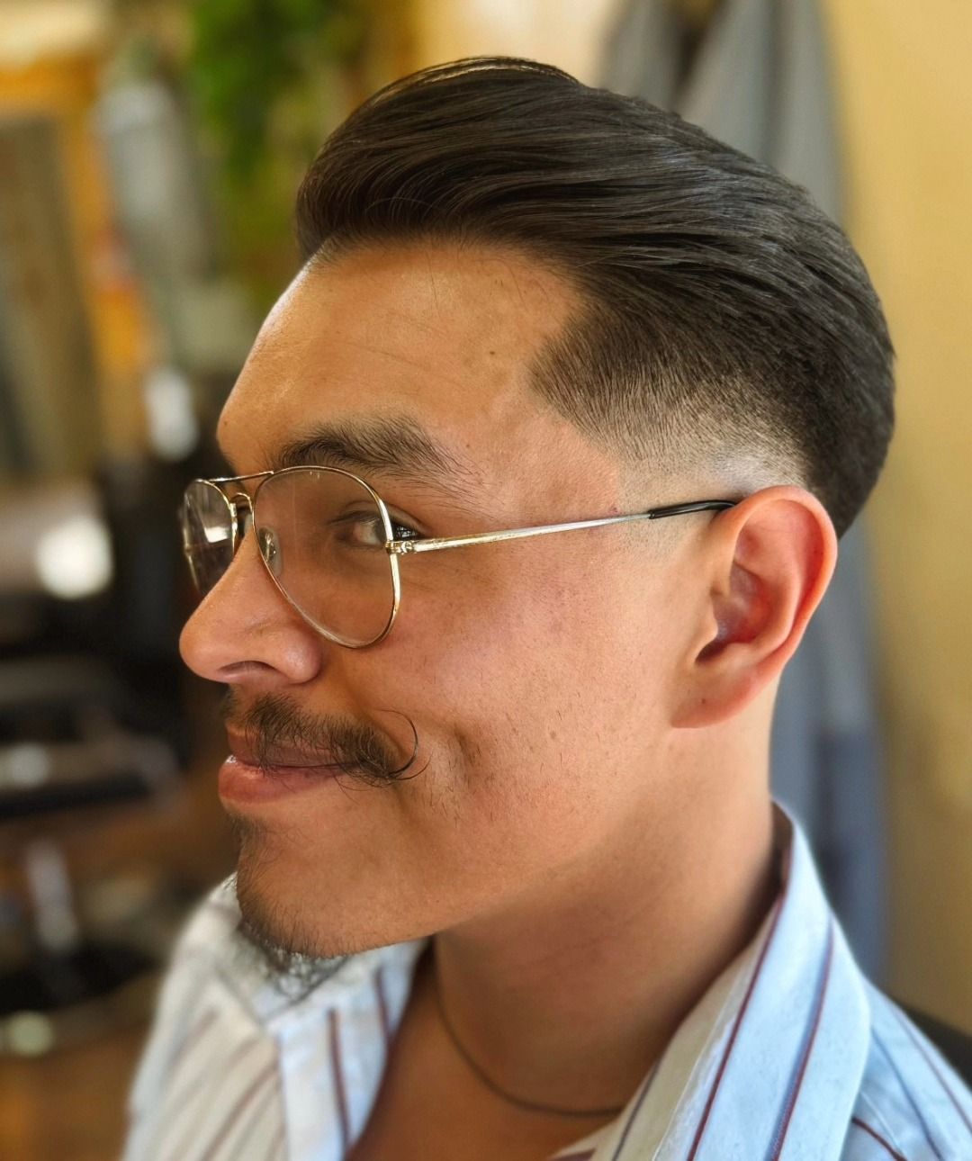 Side-profile portrait of a man with glasses and a mustache, in a light blue striped shirt, indoors with a blurred background