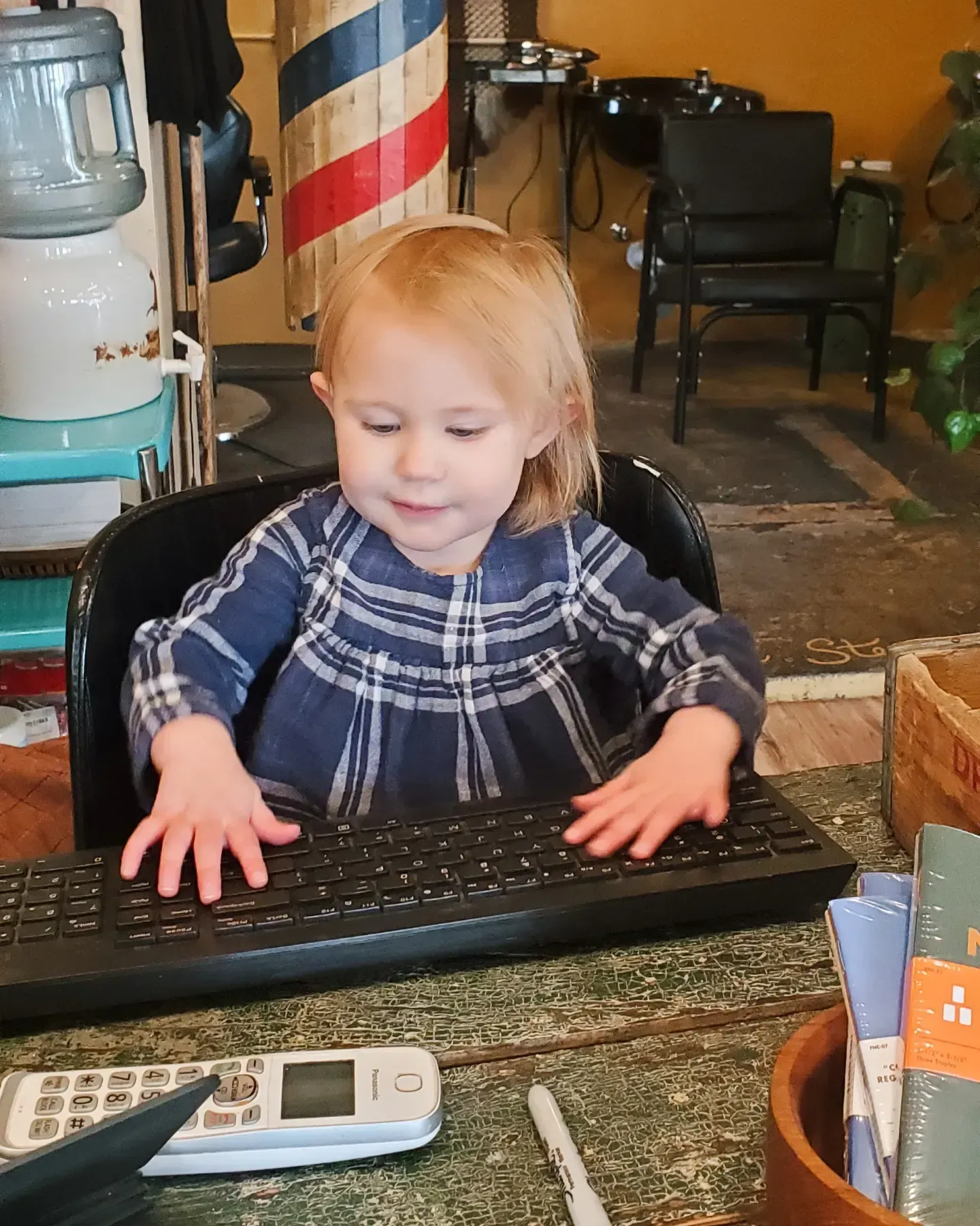 Toddler at a desk typing on a black keyboard, with a phone nearby in a cozy room.