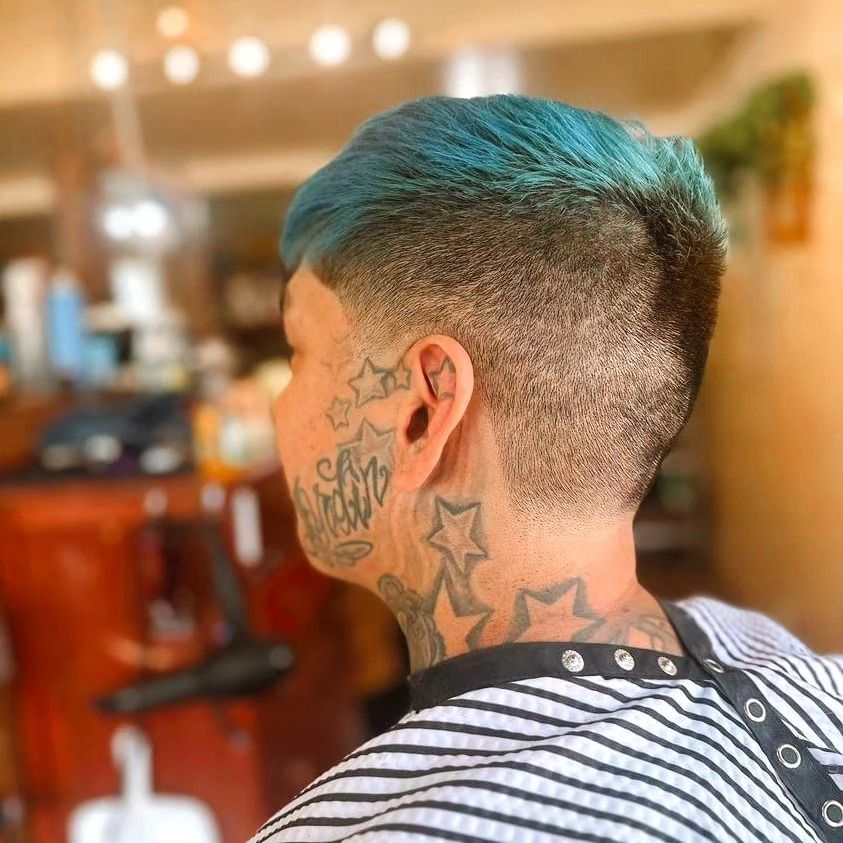 Person with teal hair and facial tattoos in a striped shirt, seen from behind in a barbershop
