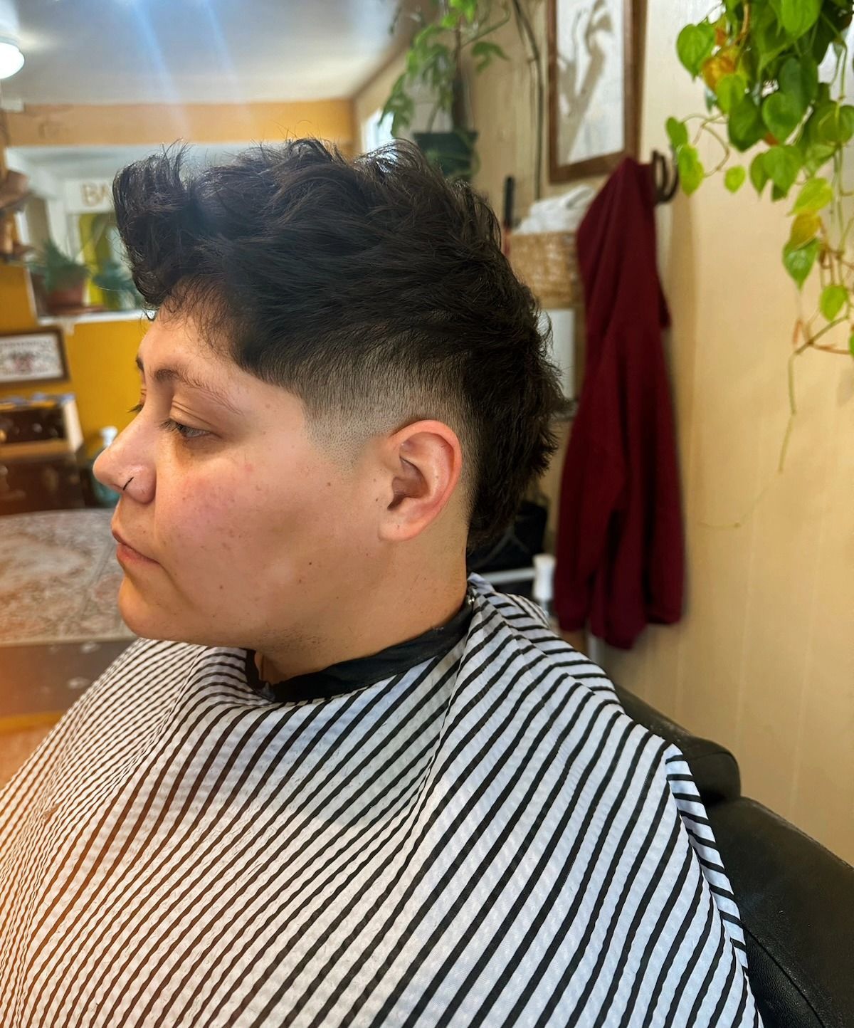 Profile view of a person with short dark hair in a striped barber cape, seated in a salon chair.