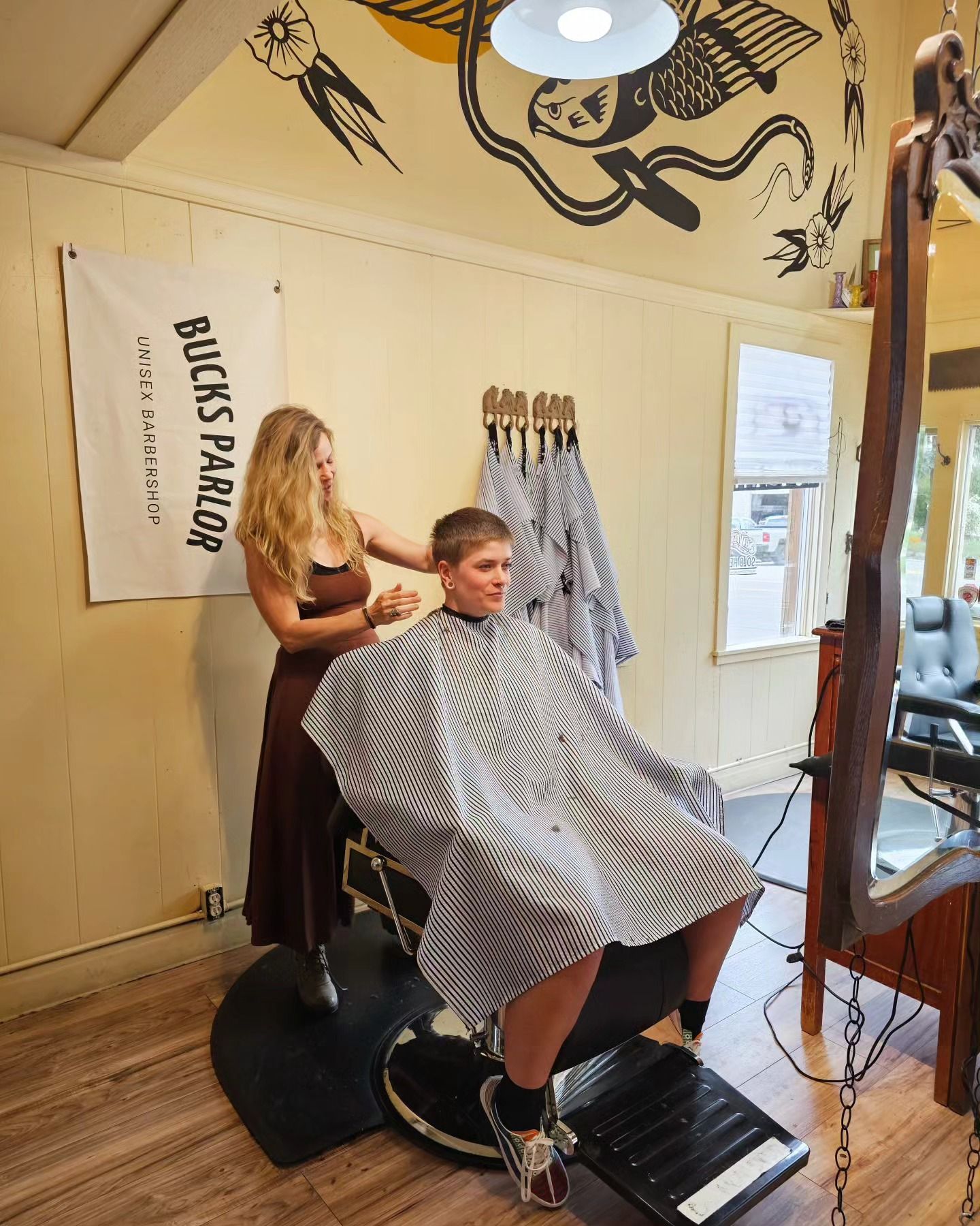 Haircut in progress at a salon, with a client under a gray cape and stylist standing behind.