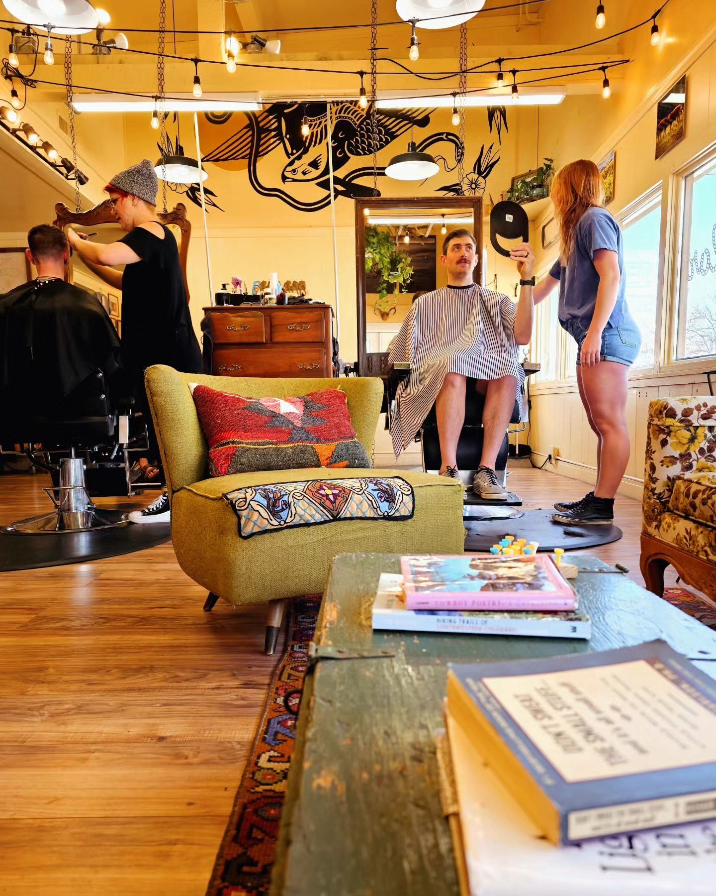 People standing in a cozy living room with warm lighting, a green chair, and a coffee table with books.