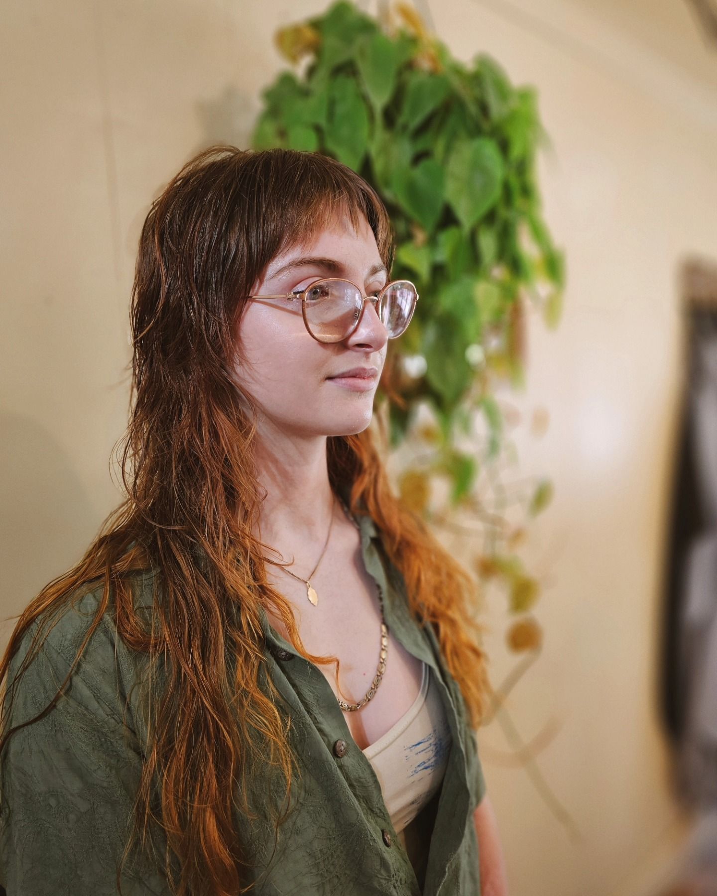 Woman with glasses and wet hair in a green shirt, standing indoors near a leafy plant.