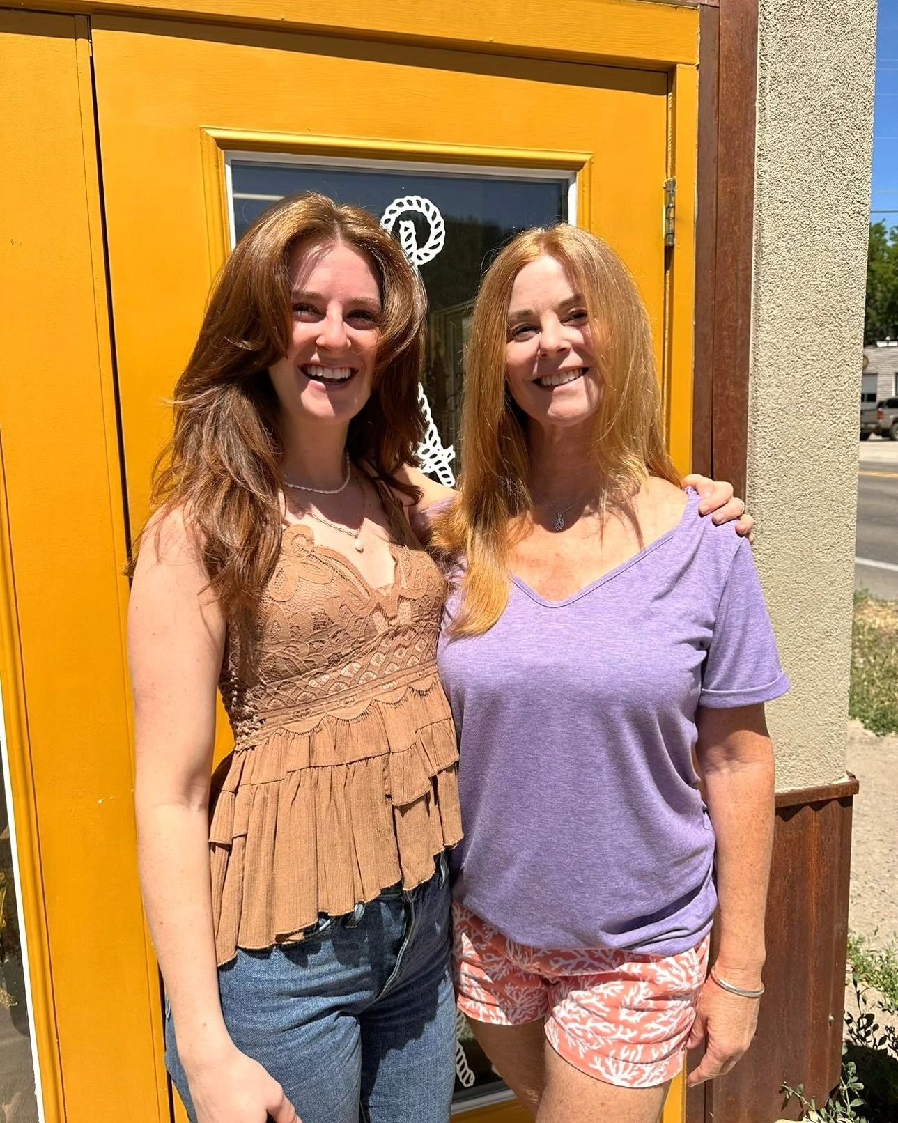 Two women smiling outdoors by a yellow door in bright sunlight.