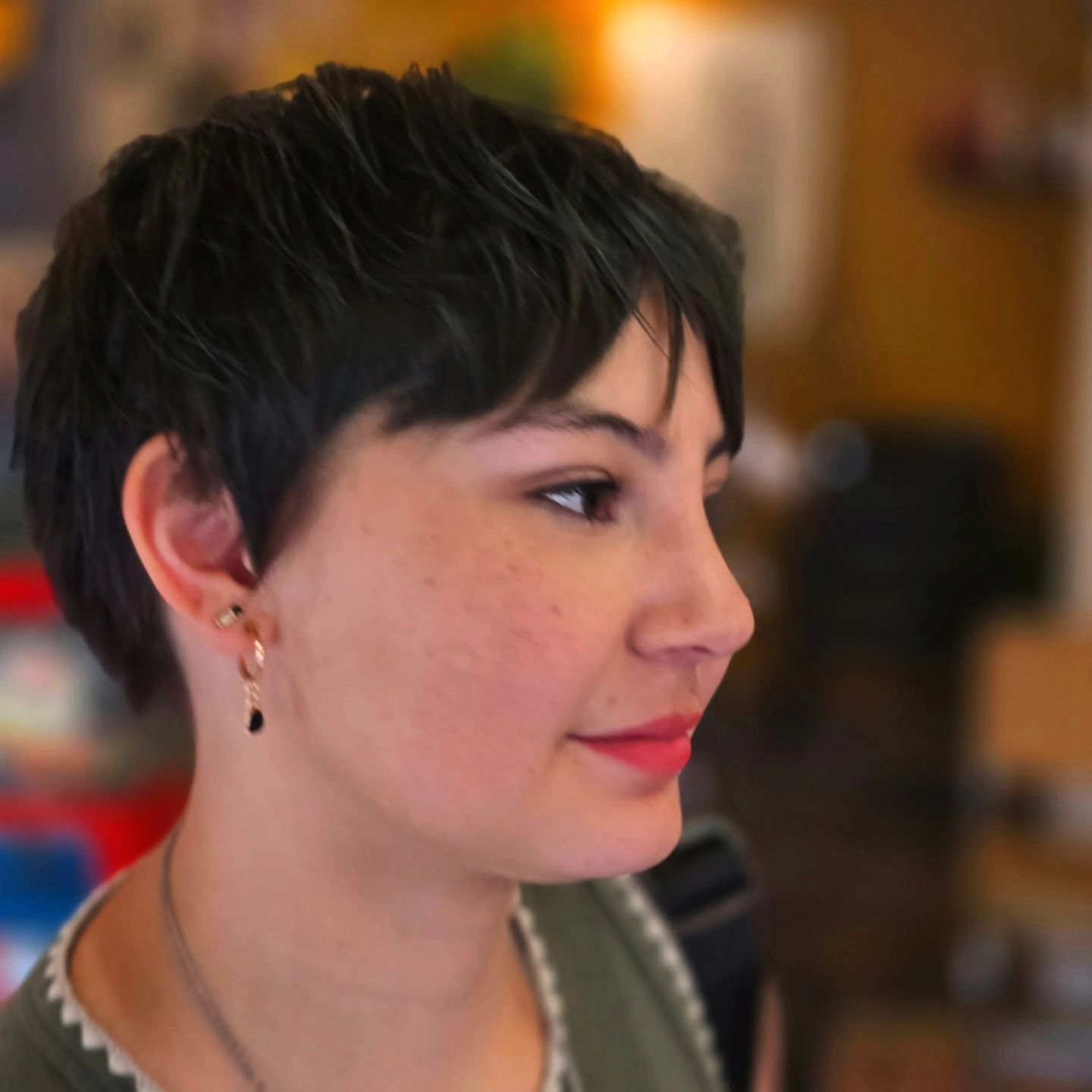 Profile view of a person with short dark hair and earrings, indoors with a softly blurred warm background