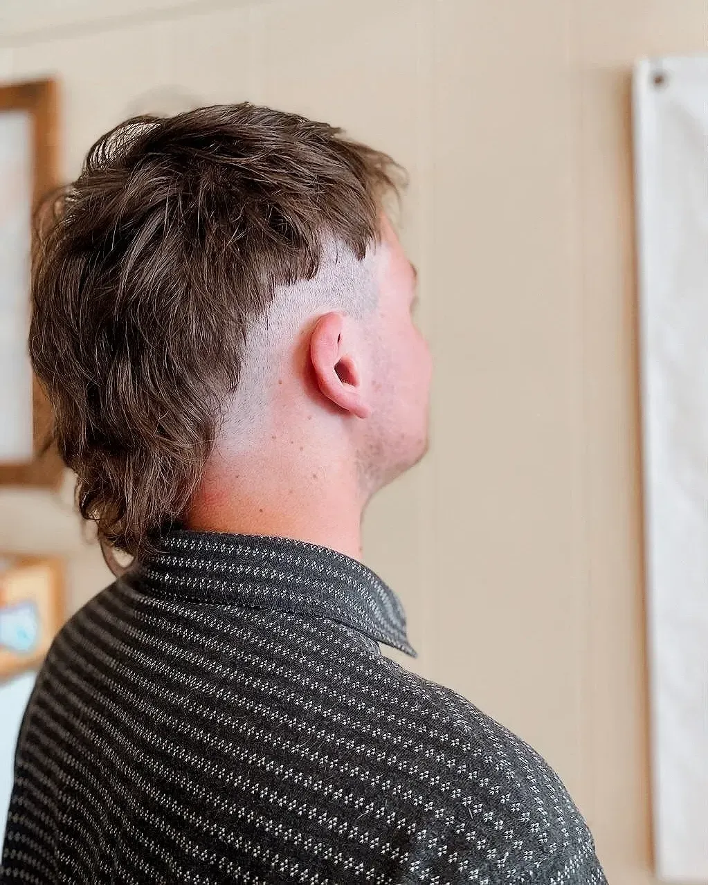 Person seen from behind with a short mullet haircut, wearing a dark patterned shirt indoors.