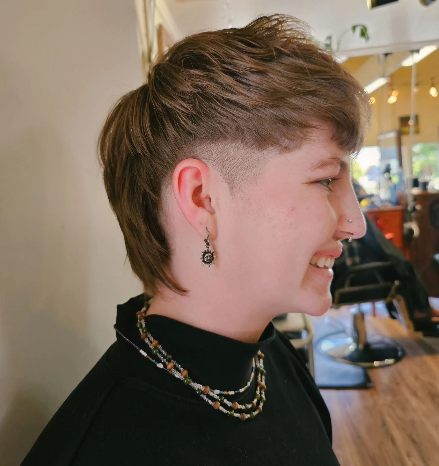 Profile of a smiling person with short brown hair and earrings, wearing a black top in a bright indoor space