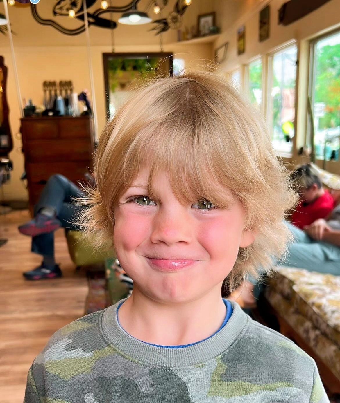 Smiling child indoors with blond hair, wearing a camouflage shirt, in a cozy room with people in the background