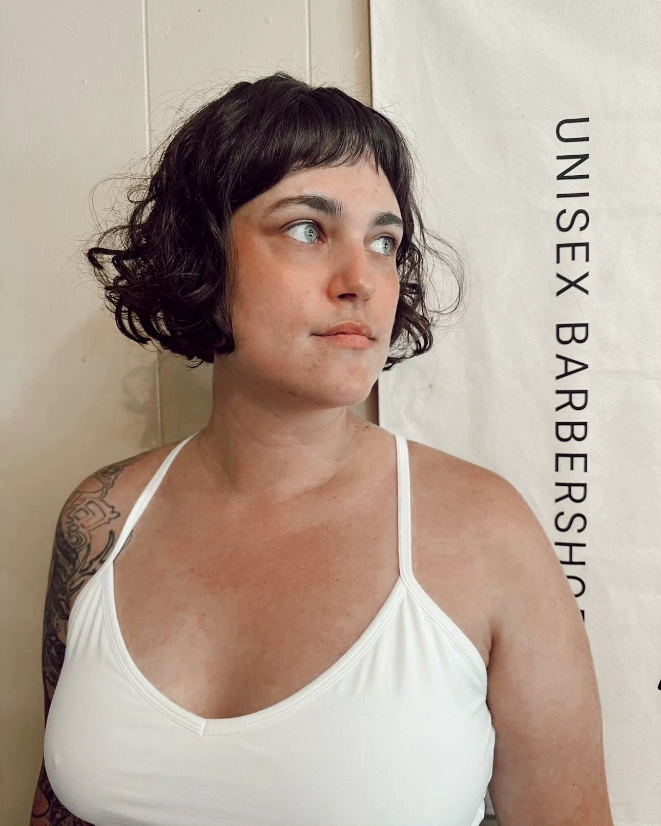 Woman with short dark hair in a white top beside a sign reading “UNISEX BARBERSHOP”