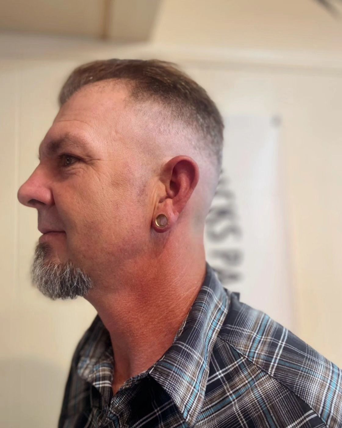 Profile view of a man with a short gray beard, wearing a plaid shirt and earring, indoors.
