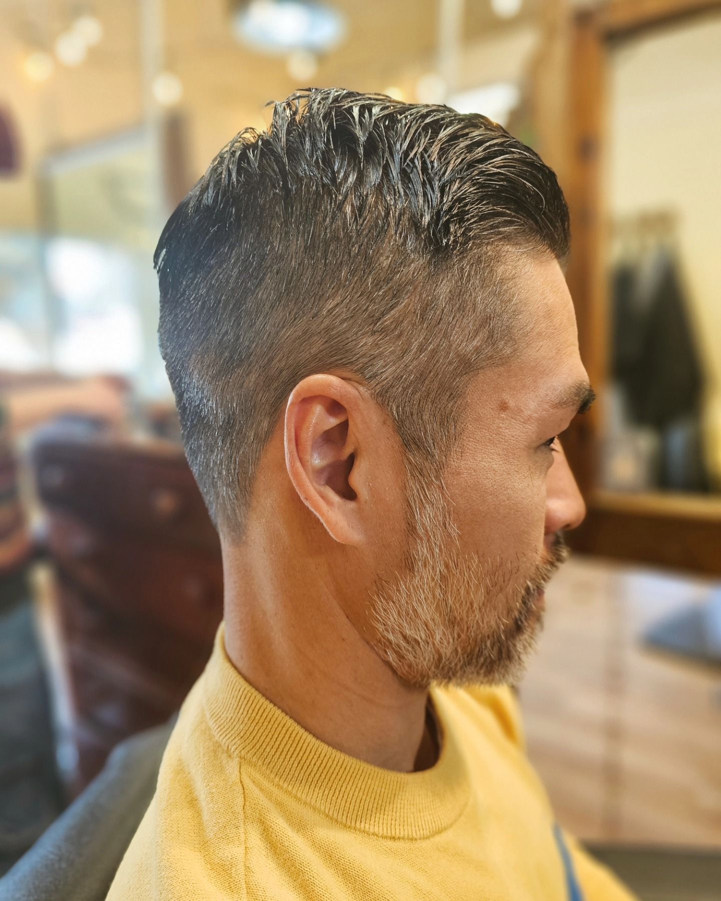 Side profile of a man with a short fade haircut and beard, wearing a yellow shirt, in a barbershop.