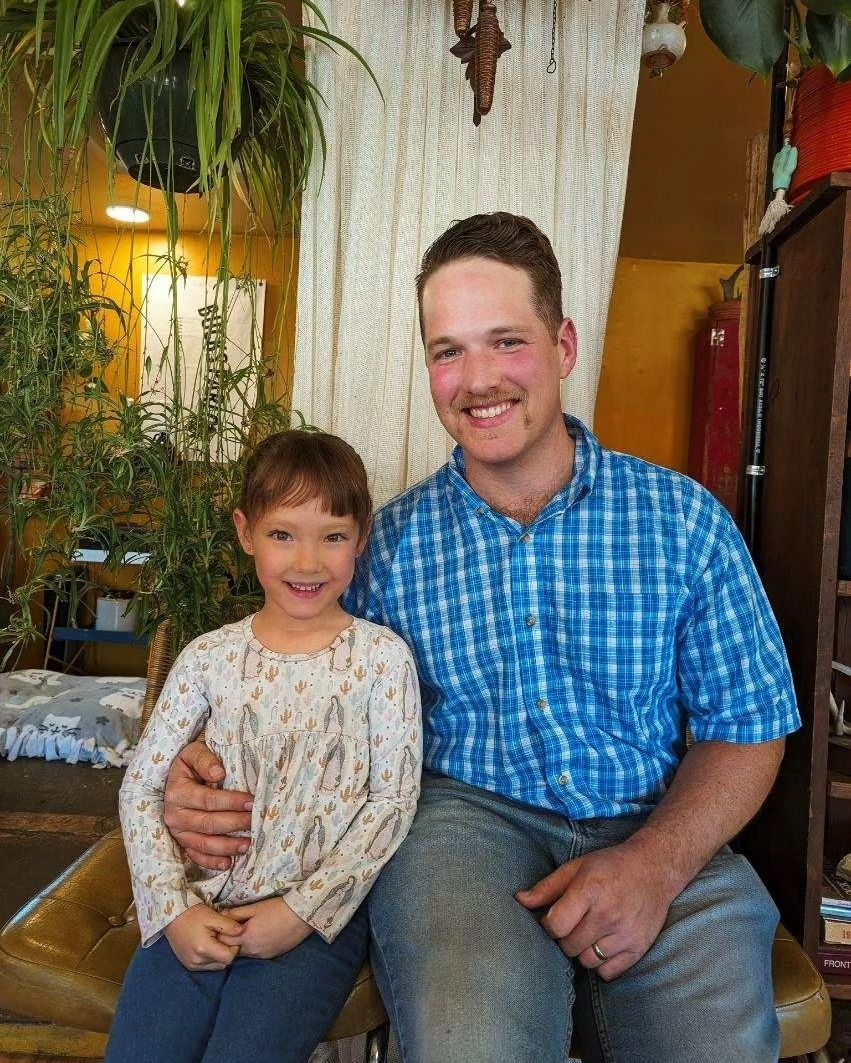 Smiling adult and child sitting together on a bench indoors, with plants and warm lighting behind them