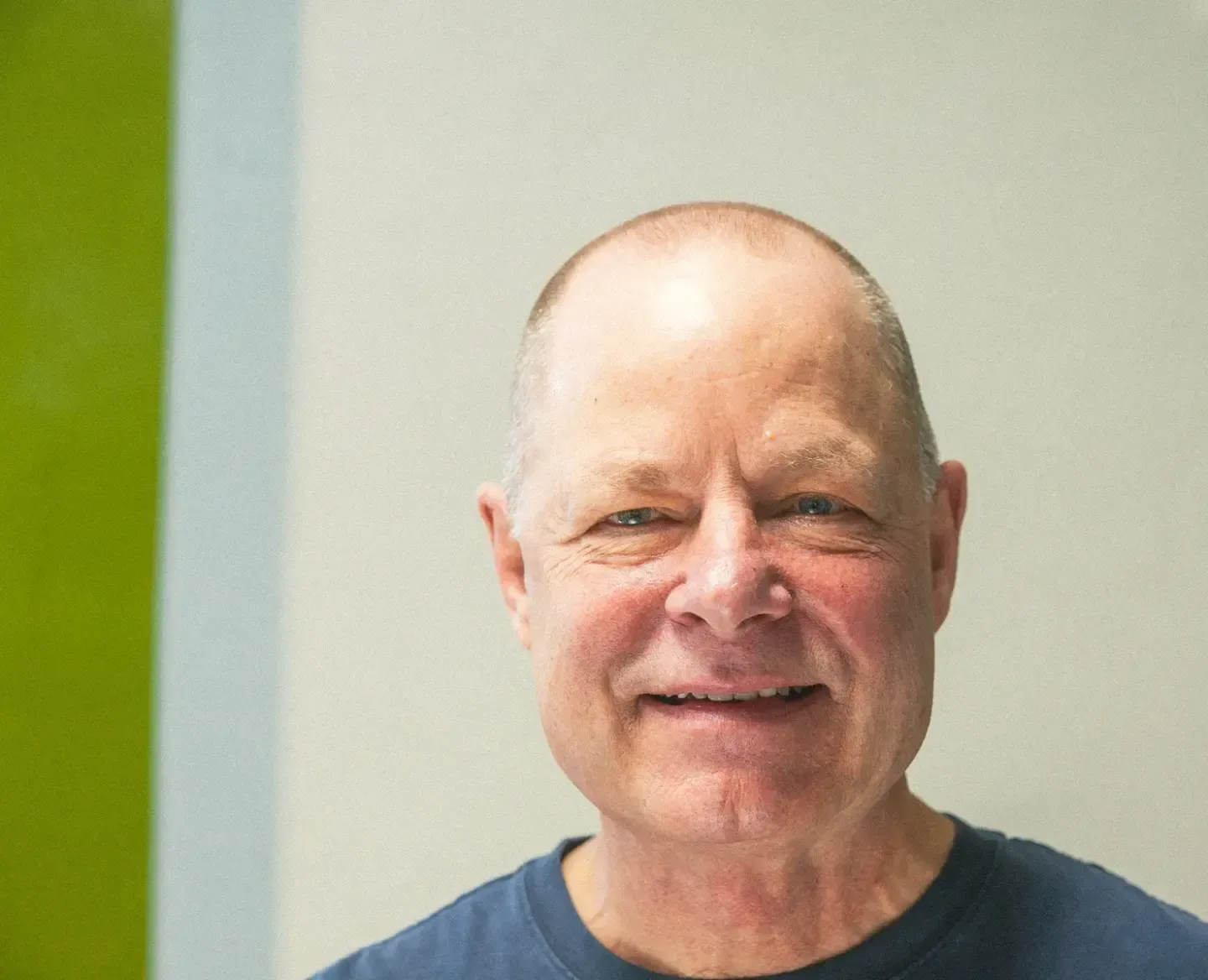 Smiling bald man in a blue shirt against a light wall with a green strip on the left