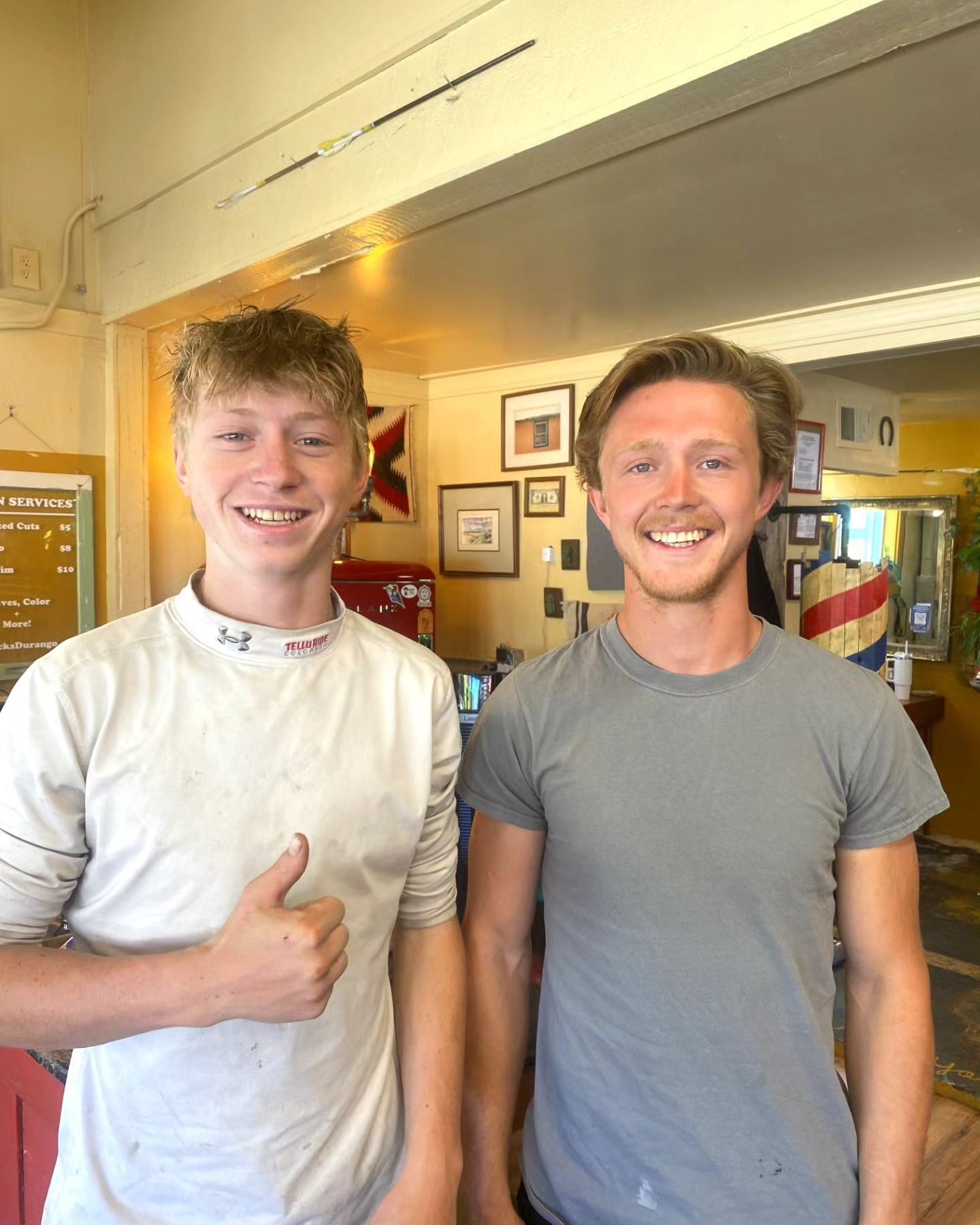 Two smiling people standing indoors, one giving a thumbs-up, in a bright café or shop.