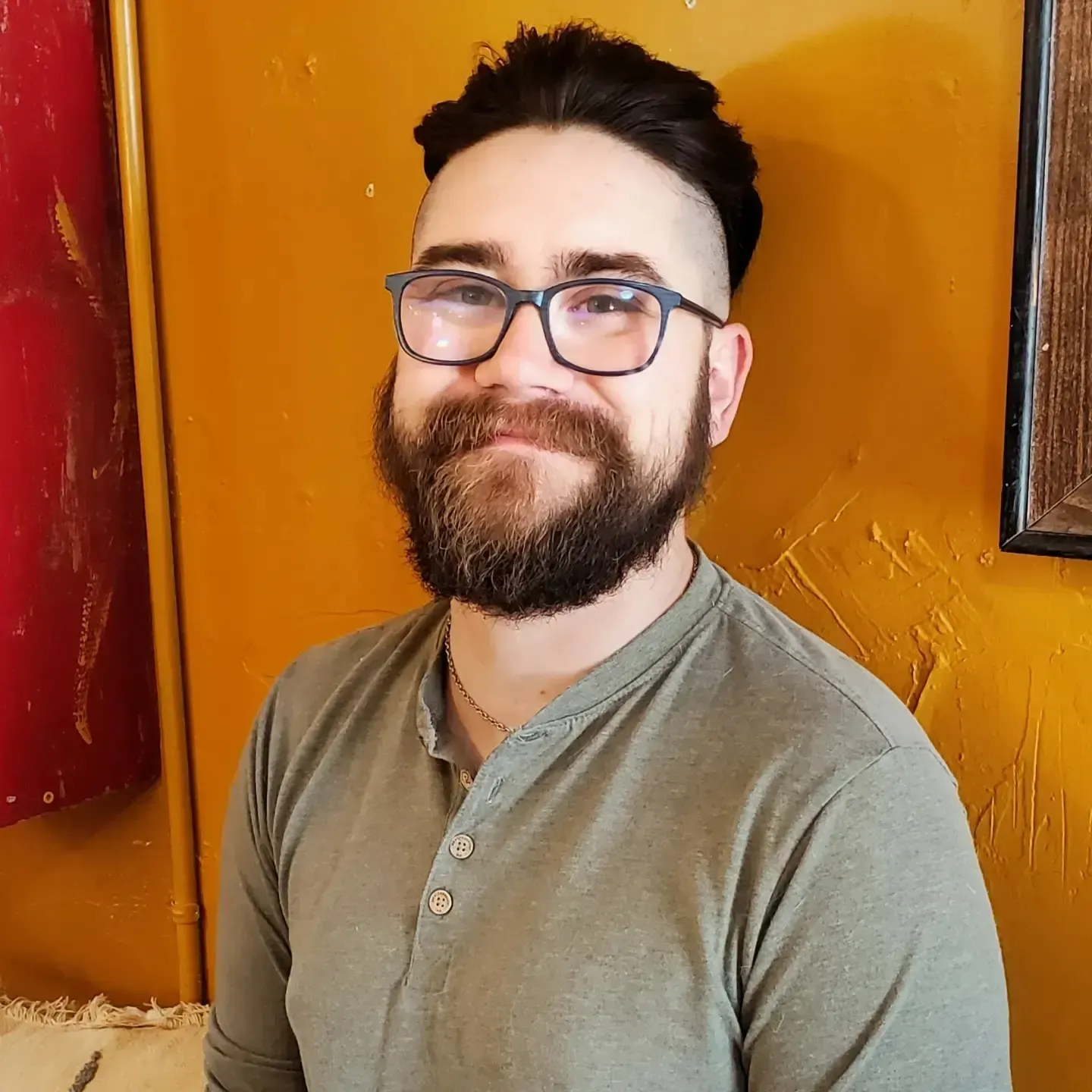 Smiling bearded person wearing glasses and a gray shirt, standing against an orange wall.