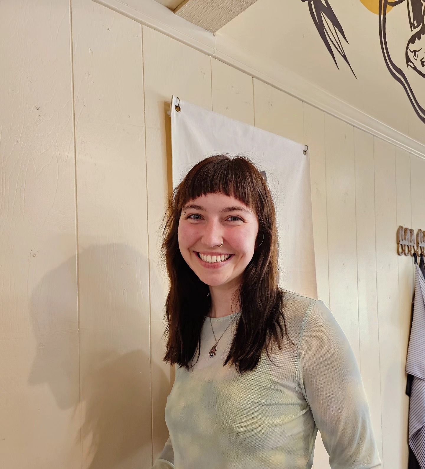 Smiling person with dark hair in a light top standing indoors against a beige wall.