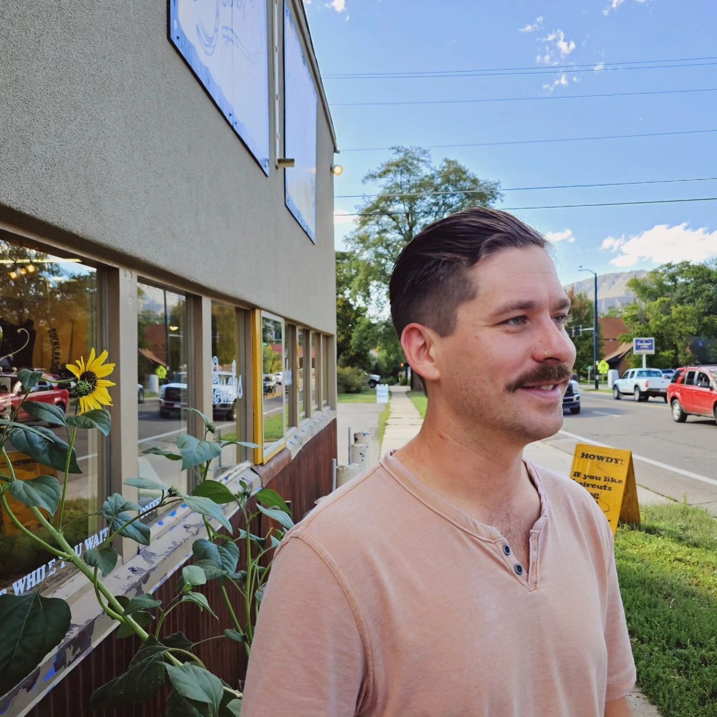 Man with mustache standing beside a building on a sunny sidewalk with flowers in the window.