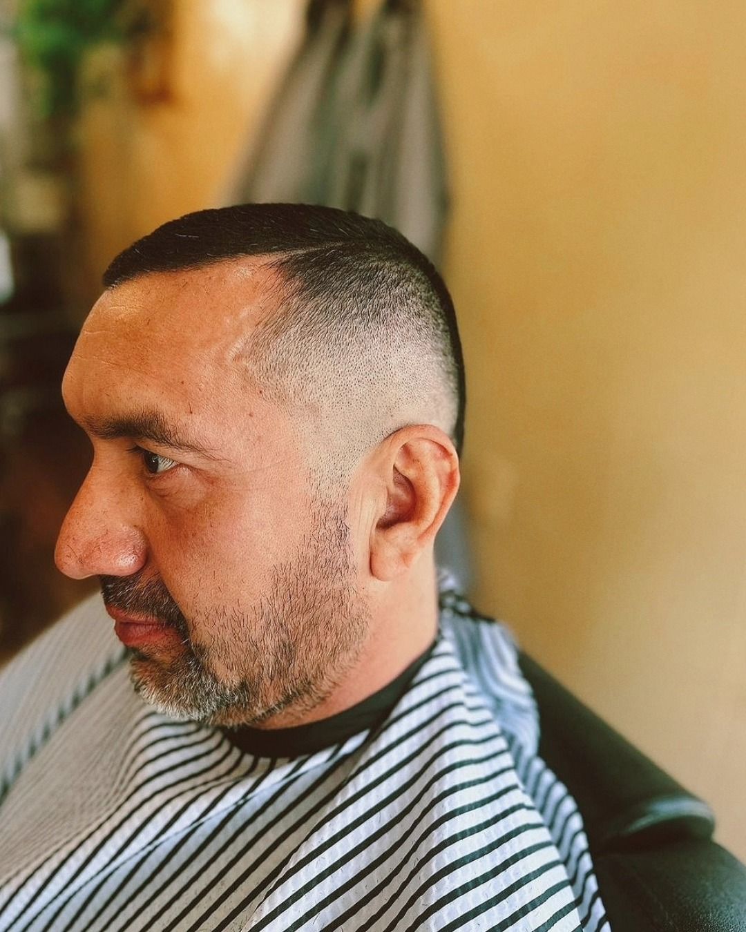 Side-profile of a man with a close-cropped haircut and beard, wearing a striped barber cape.