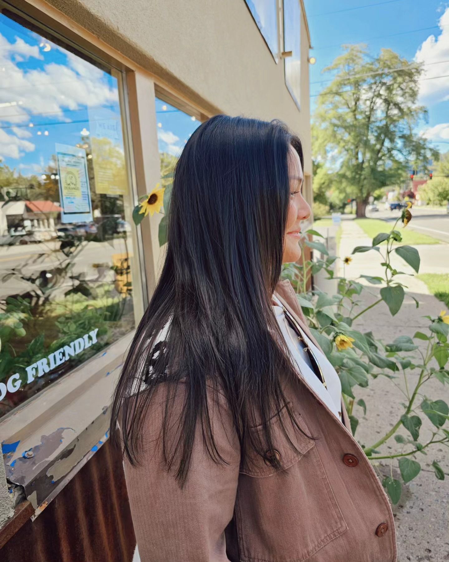 Woman with long dark hair in a brown jacket standing beside a storefront window on a sunny street