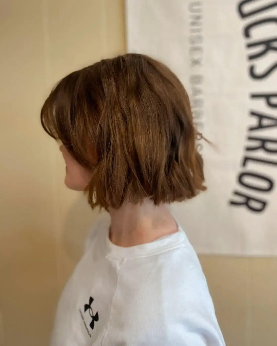 Profile view of a person with short brown hair wearing a white T-shirt against a tan wall and sign backdrop