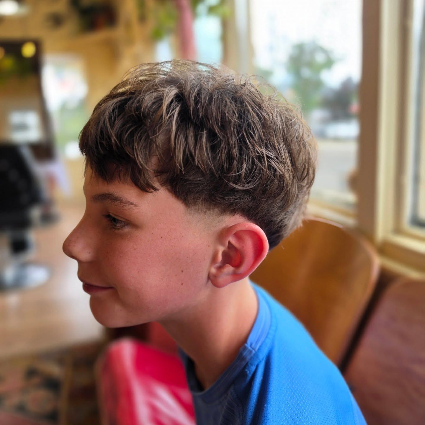 Profile of a child with short brown hair in a blue shirt, seated indoors by a window