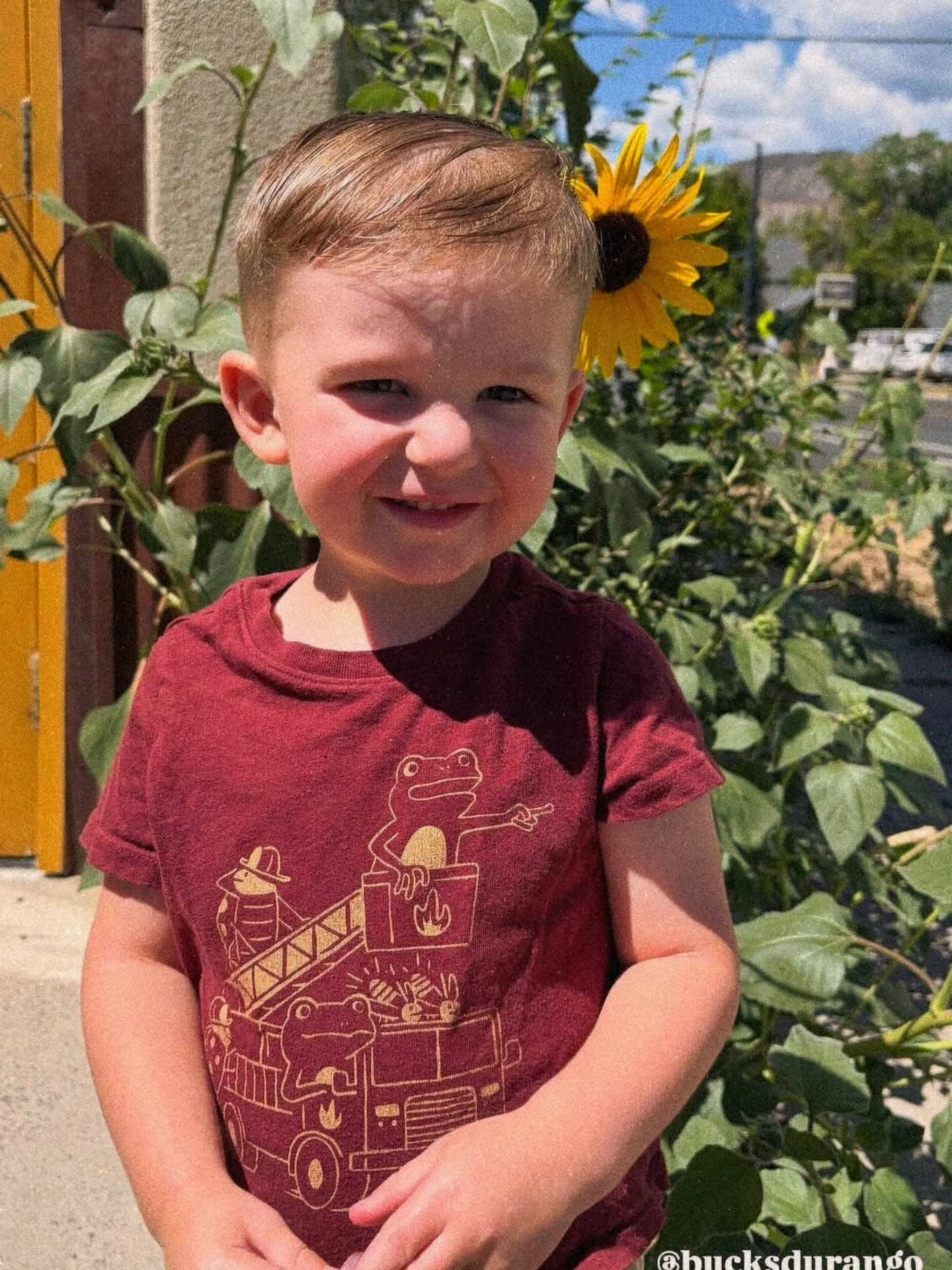 Young child in a red shirt standing outdoors in front of a sunflower and garden greenery